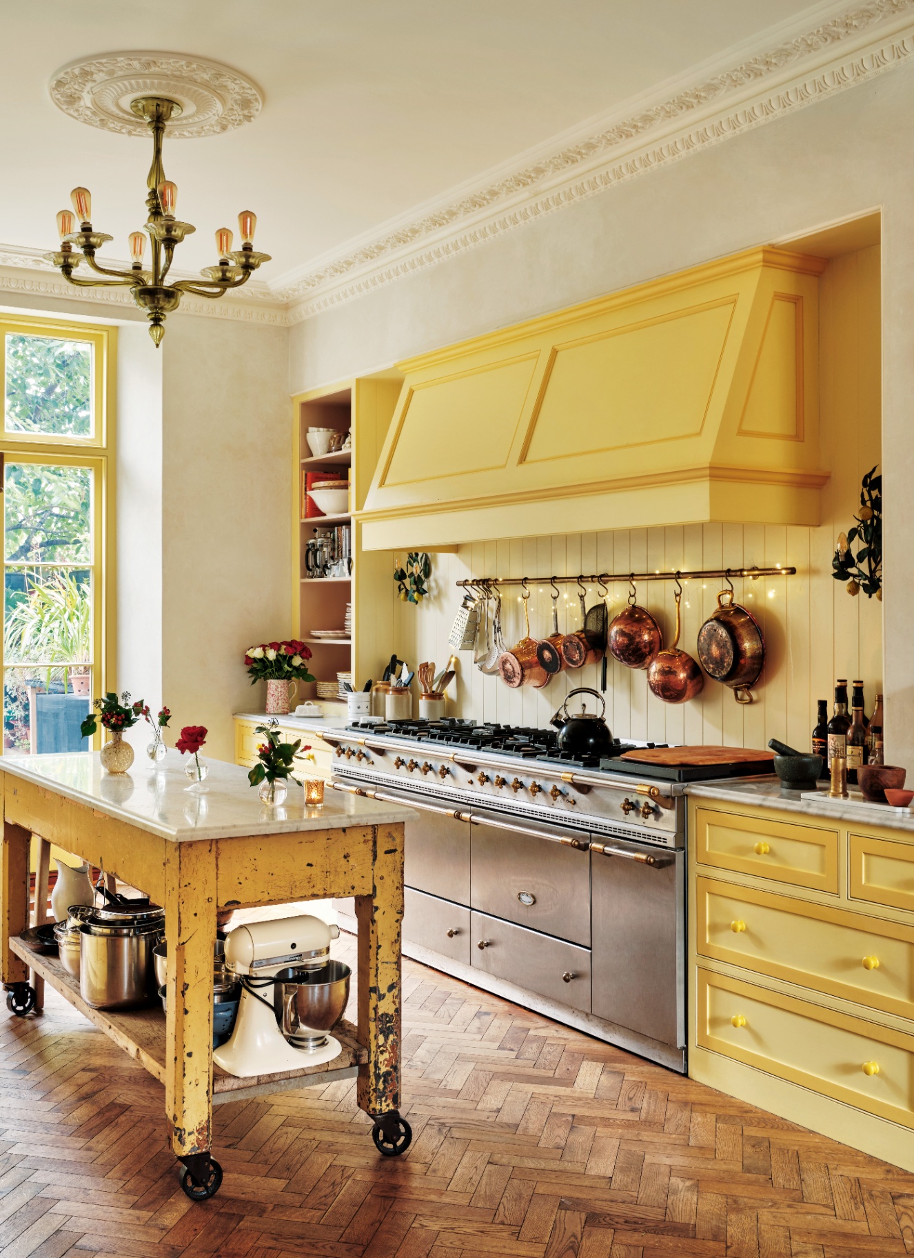 a kitchen with bright yellow cabinetry, a yellow kitchen island, a large silver range cooker and a row of copper pans hanging above the cooker