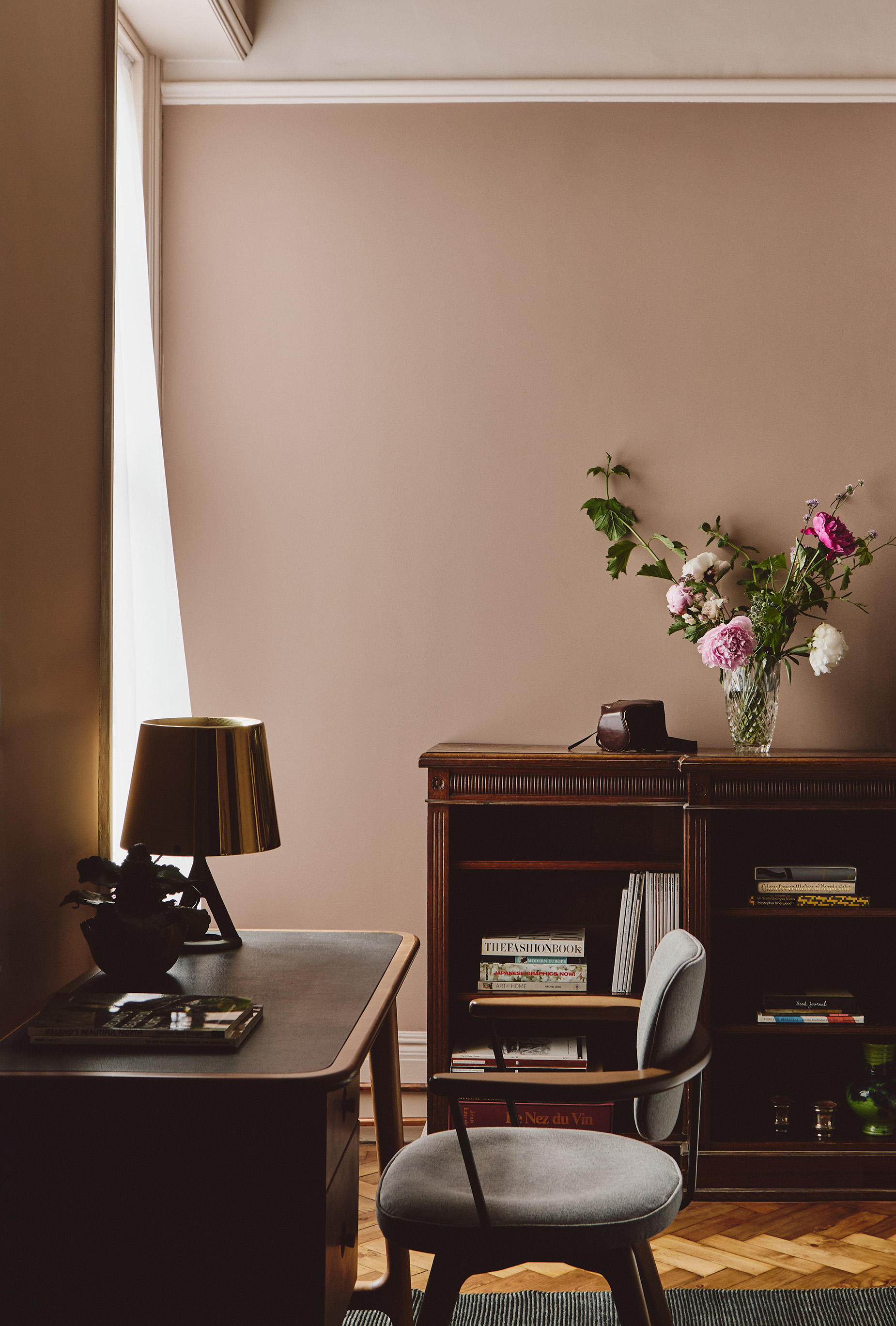  A room with warm pink walls and wood flooring. In the foreground is a wood desk with curved corners, topped with a plant and table lamp and paired with a grey chair. In the background is a half bookshelf made from wood, styled with books and flowers