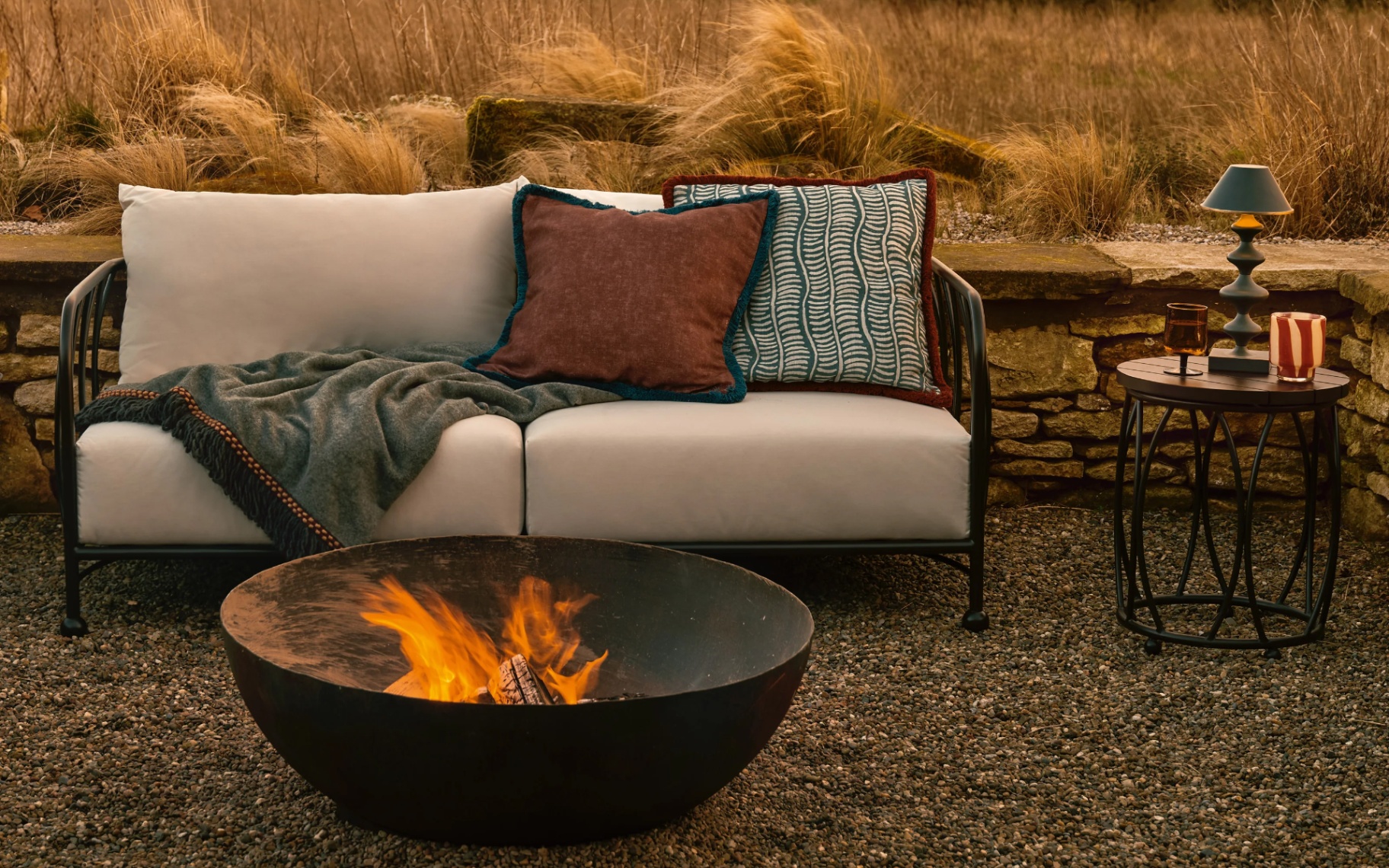 a large black fire pit with an open flame on the floor in front of an outdoor sofa with beige cushions and long dry grass in the background