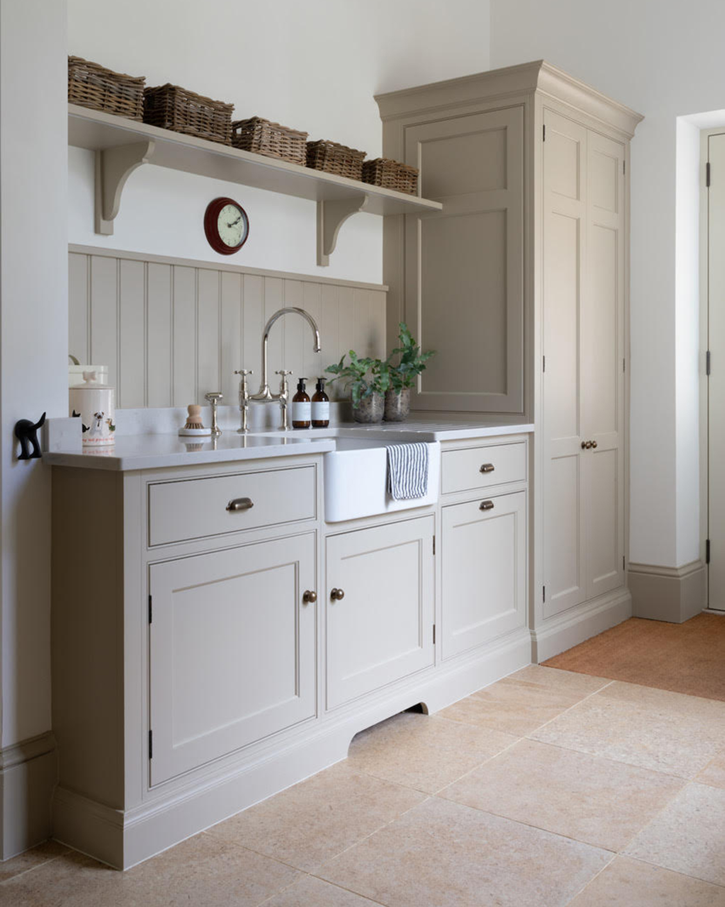 A UTILITY ROOM with cream stone floors, pale beige cabinetry, a butlers sink and an open shelf above it with wicker baskets