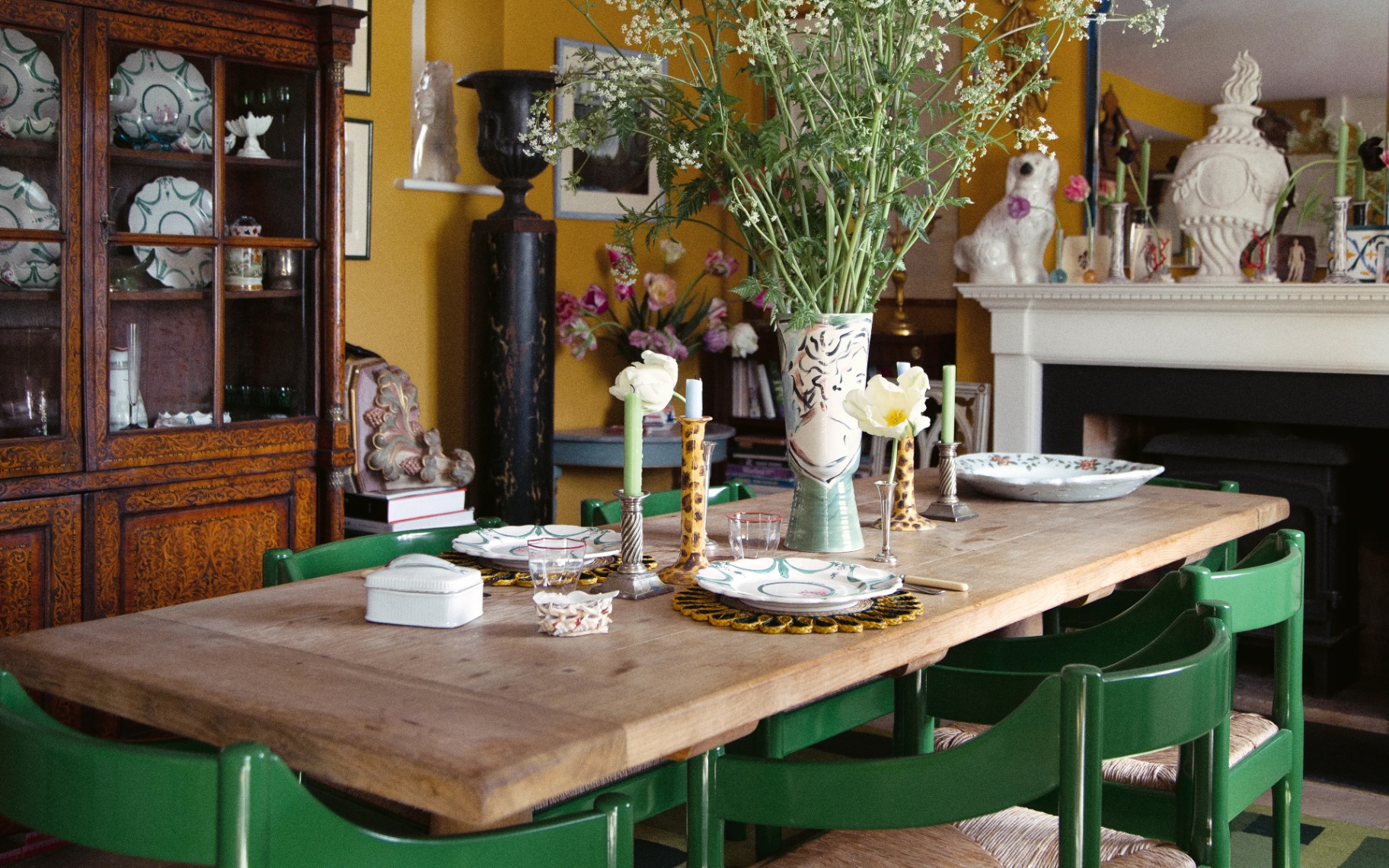 Mustard yellow dining room featuring a long wooden table surrounded by green chairs, a tall floral vase centerpiece, a large vintage display cabinet, and a white fireplace topped with a blue-framed mirror