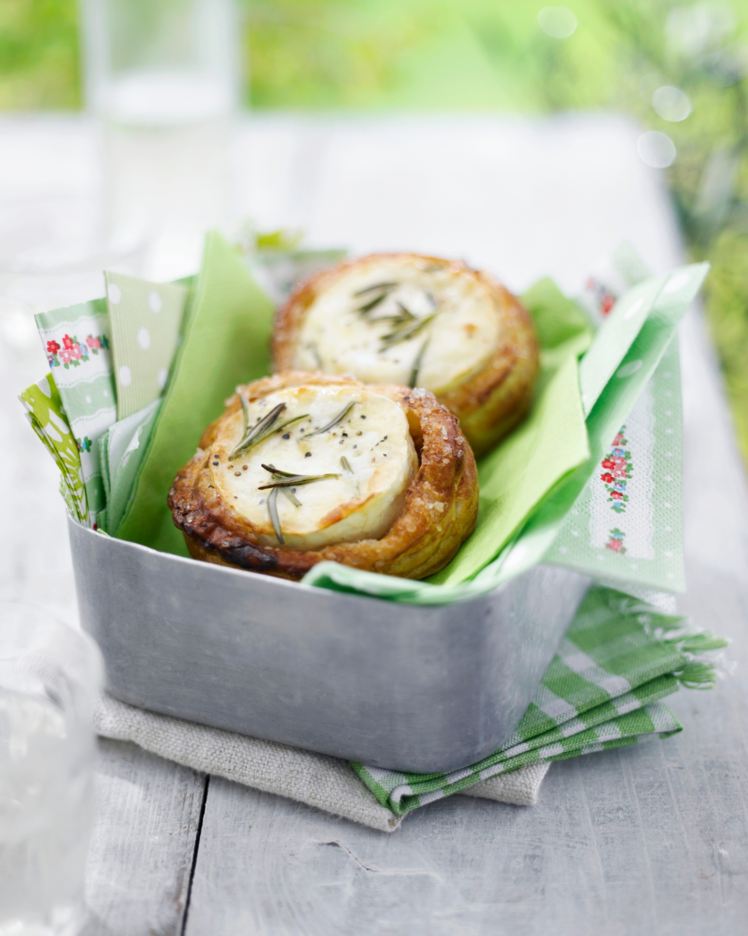 two savoury pastry tarlets with goats cheese in a metal container on top of green gingham napkins on a grey wooden table