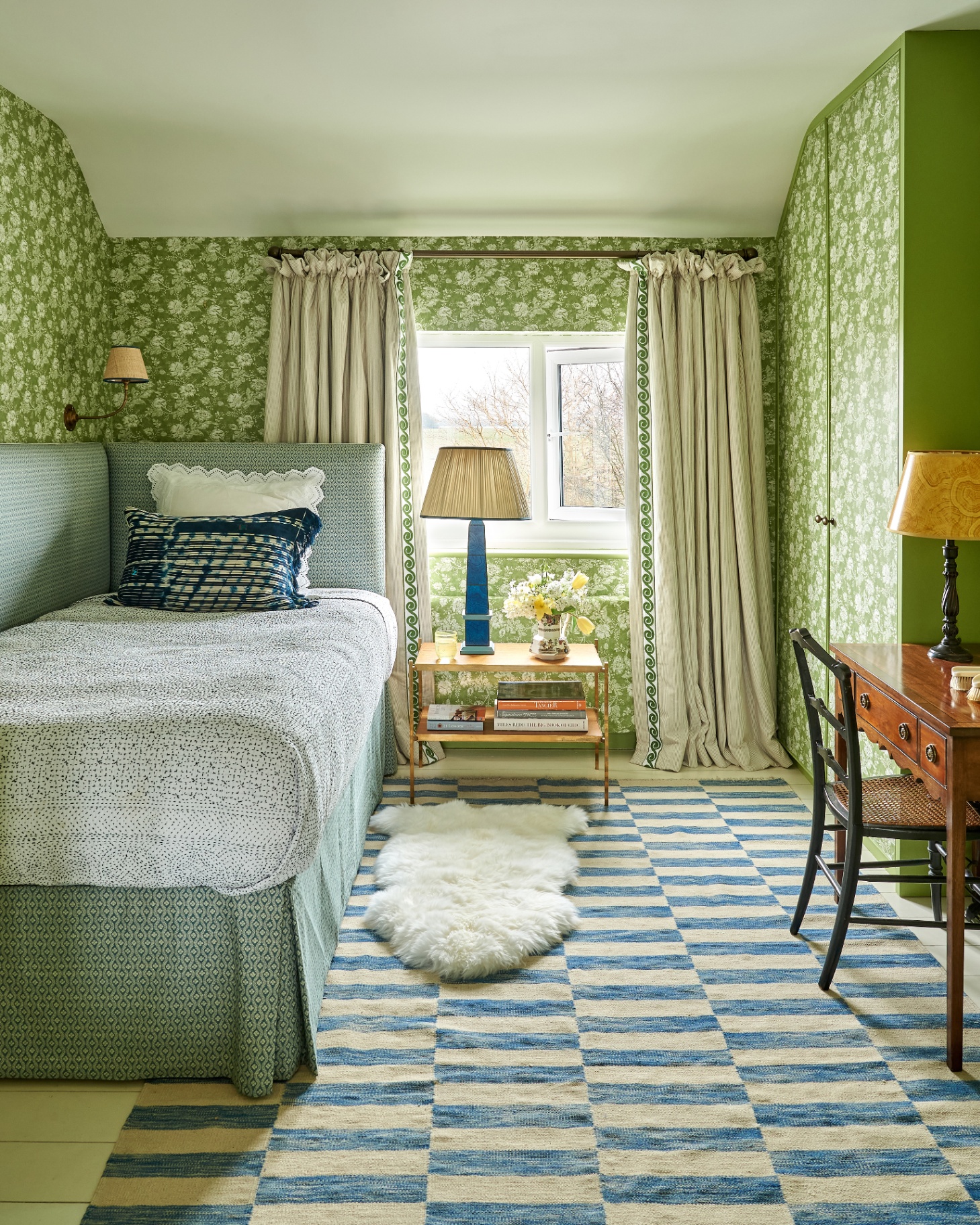 a bedroom with green floral wallpaper, a blue and cream checkerboard rug and a single bed with white bedding and a green patterned valance