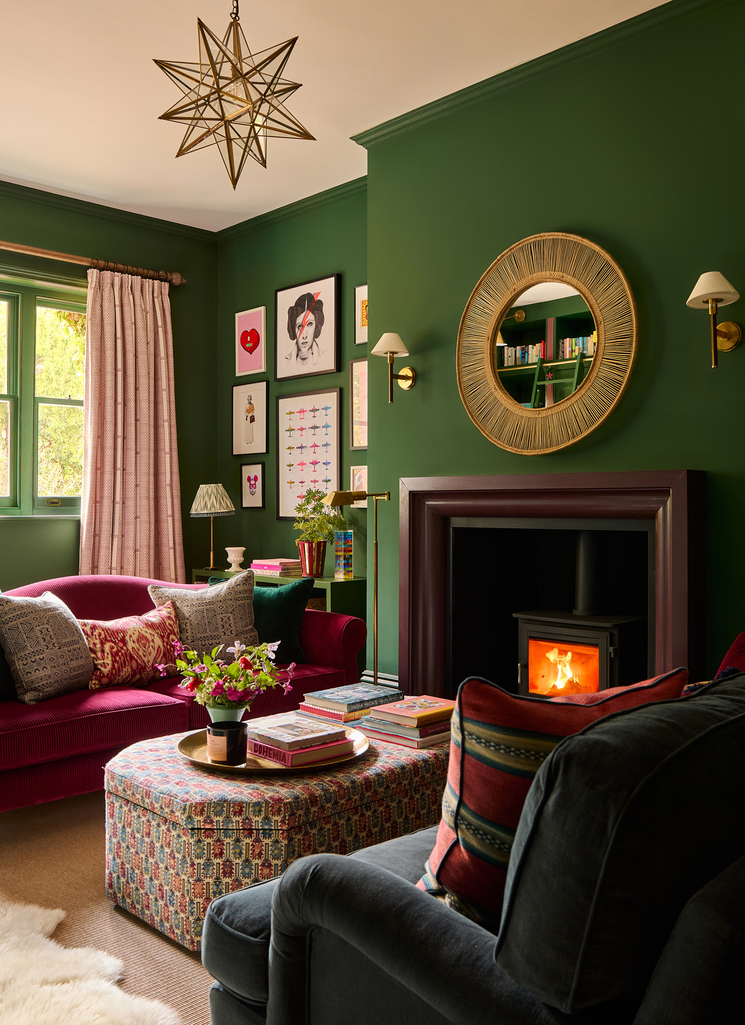 A cosy living room with rich forest green walls, a deep berry-red sofa, a starburst pendant light, and a circular gold mirror above a wood-burning stove