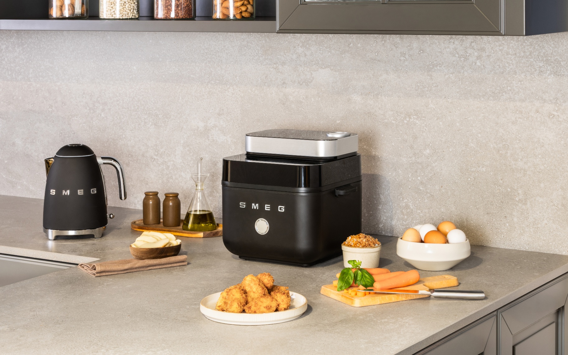 Matte black SMEG air fryer sitting on a grey stone kitchen counter alongside a matching black SMEG kettle. Plate of golden fried bites, raw carrots on a chopping board, fresh eggs, and olive oil surround the appliances against a stone wall