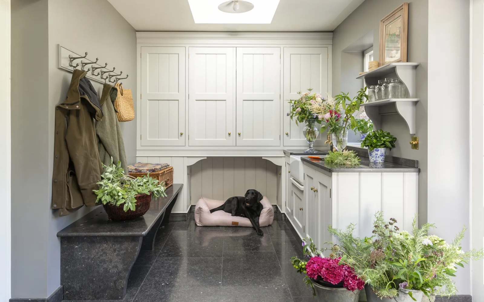 a utility room with white cabinetry, a grey granite floor, a dog bed with a black labrador inside and lots of vases full of colourful flowers