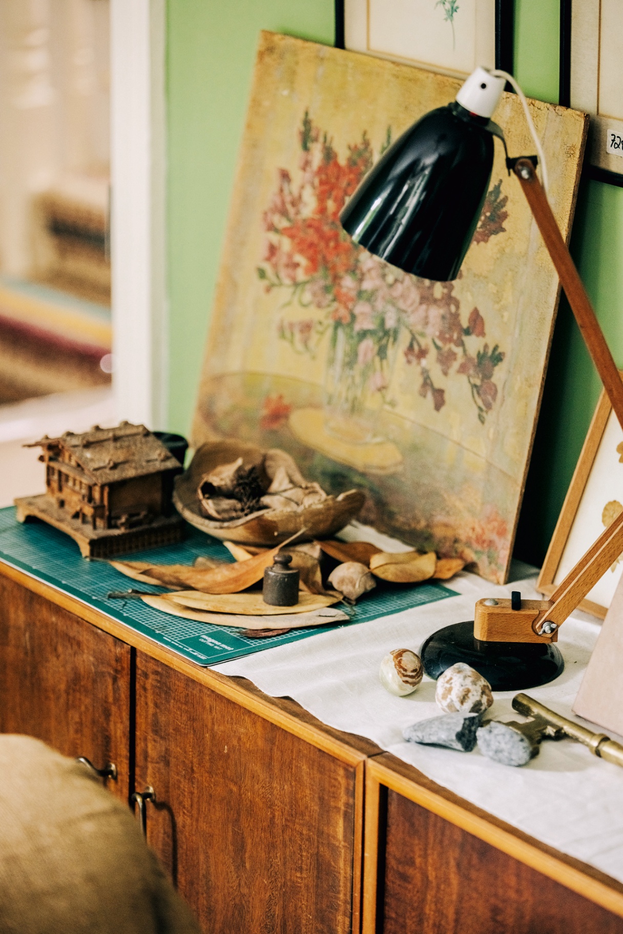 a close up of the top of a cabinet with a vintage floral artwork, wood-and-black desk lamp, leaves, stones and a wooden music box
