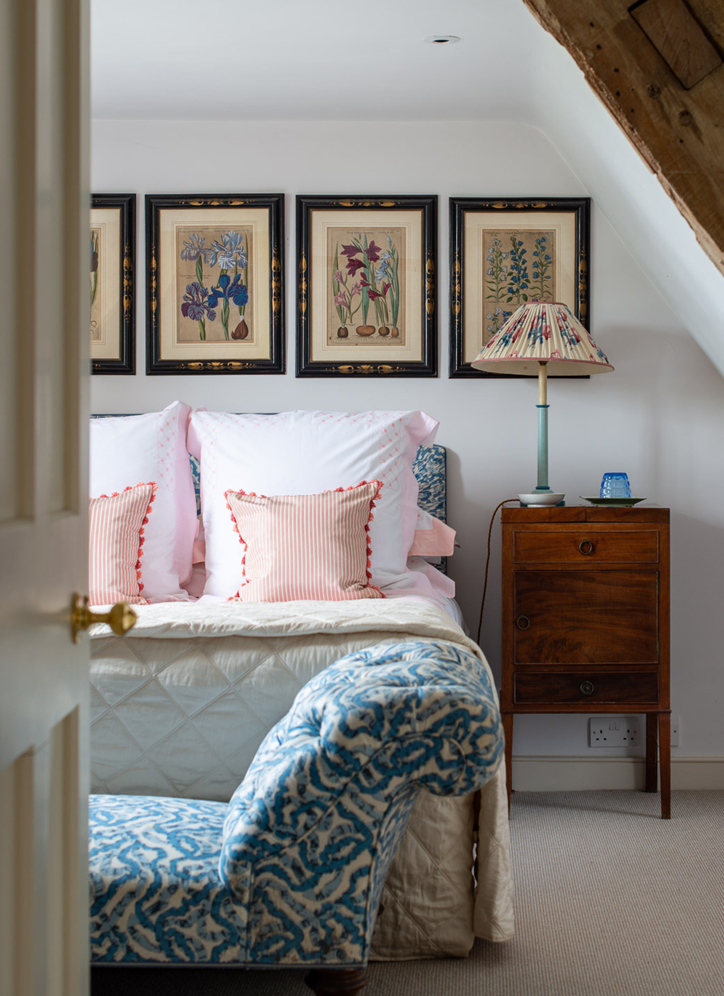 a bedroom with 4 botanical prints above the bed, a blue and white patterned ottoman at the foot of the bed and an antique bedside table