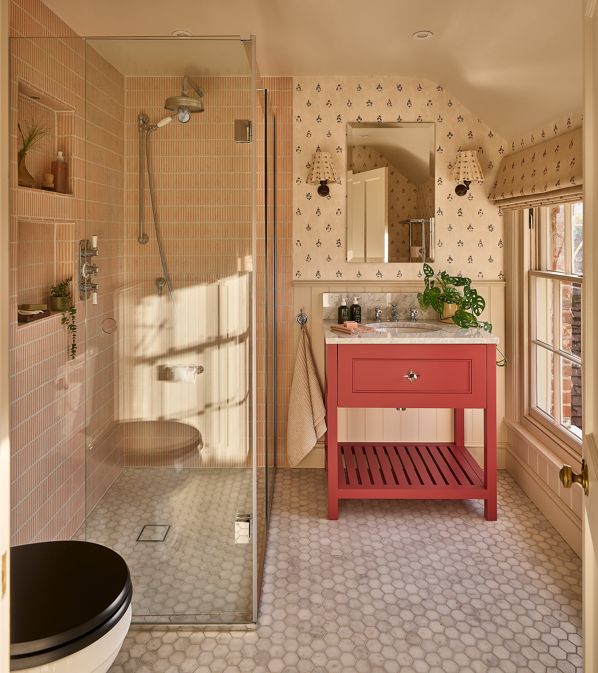 a country bathroom with a clear shower cubicle with pale pink tiles, a mosaic floor, a red vanit unit with a marble top and a swiss cheese plant on top and a rectangle mirror and two sconces above