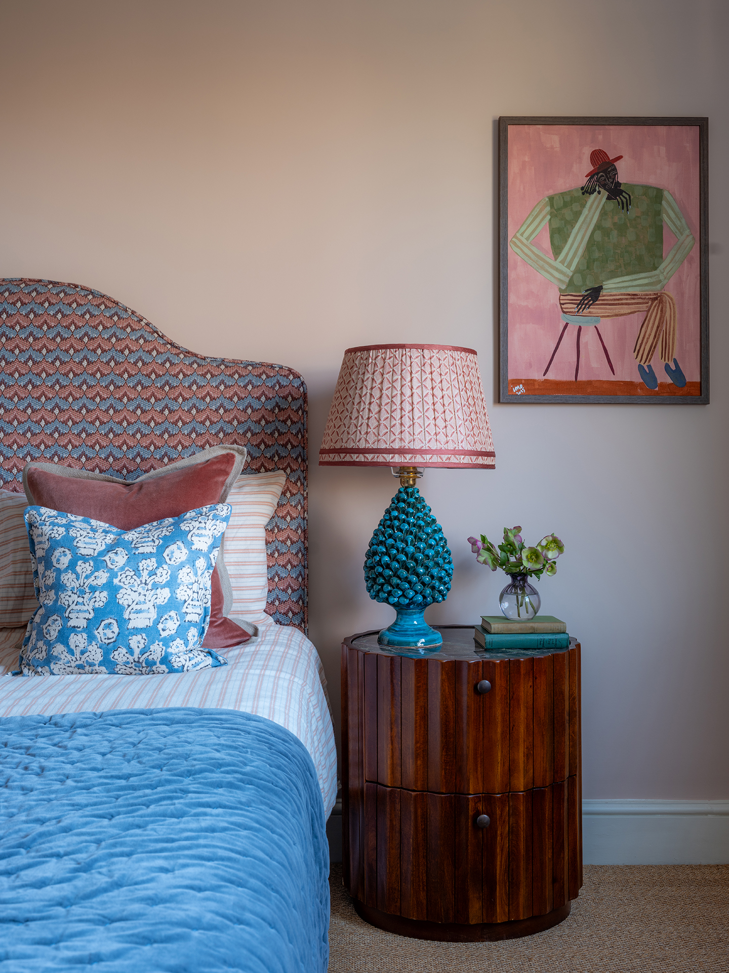 A bedroom with pink walls and a modern artwork on the wall. A bed is in the foreground with a patterned headboard and dressed with a variety of cushions and throw. A wooden fluted bedside table sits next to it, with a table lamp, books and small posey
