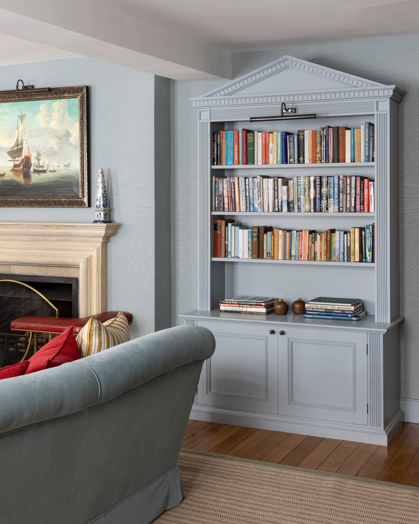 a living room corner with pale blue walls, a pale blue ornate bookcase filled with colourful books and a stone fireplace with a painting of a boat above it