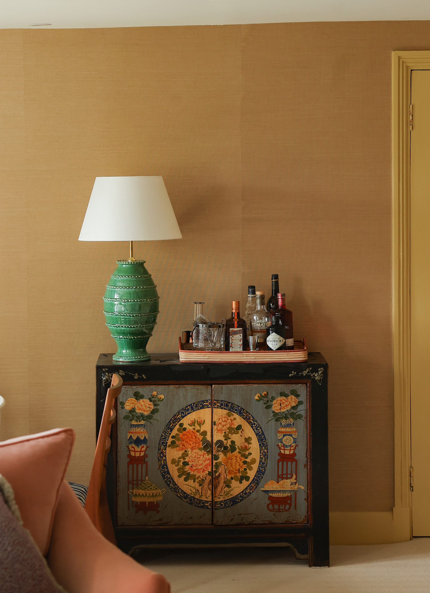 A close up of a painted decorative cabinet, topped with a green table lamp and a tray holding bottles and barware