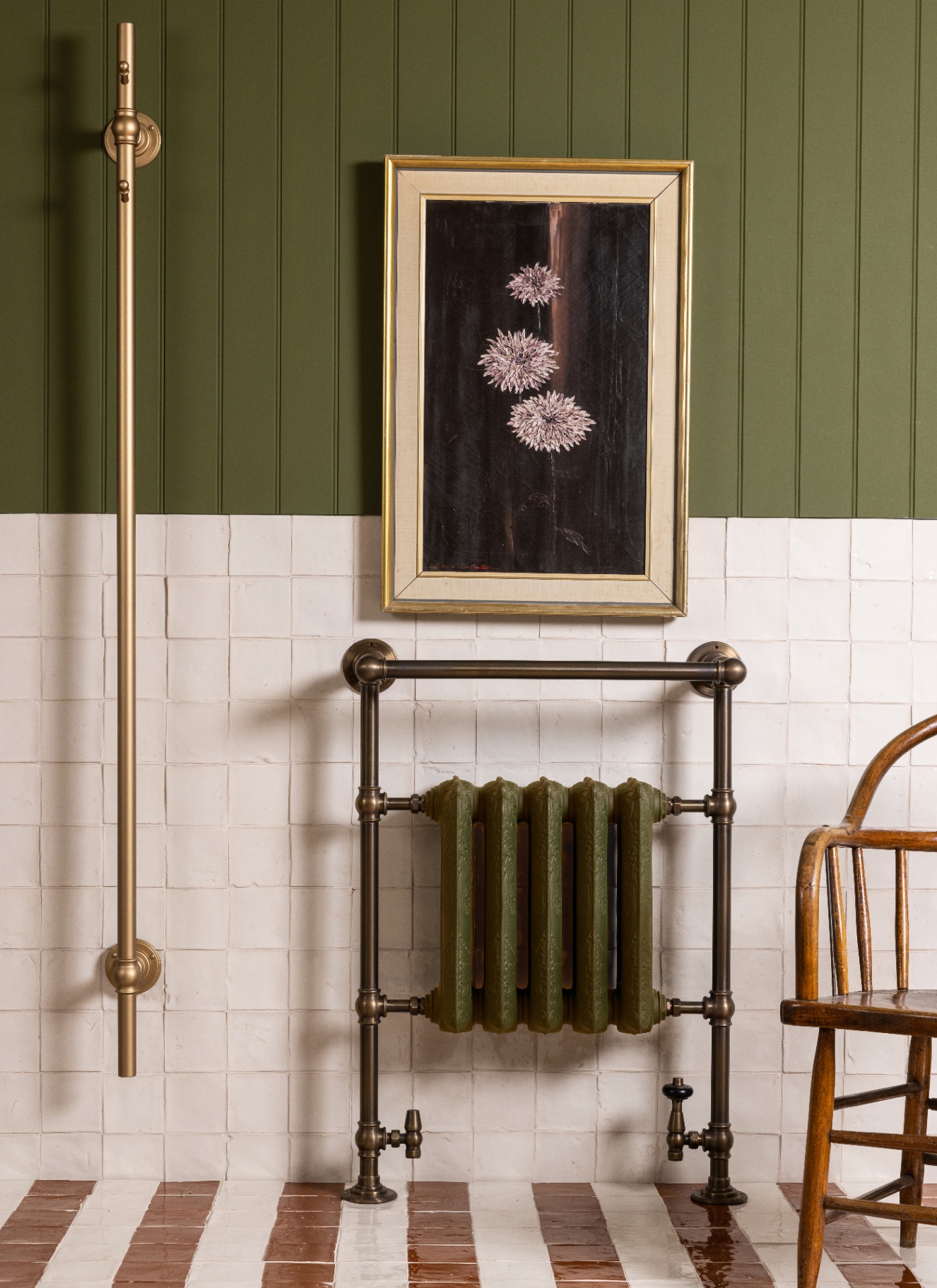 a close up of a bathroom corner with an olive green cast iron radiator, half green wall panelling, half white wall tiles, a gold framed floral painting and white and rust floor tiles