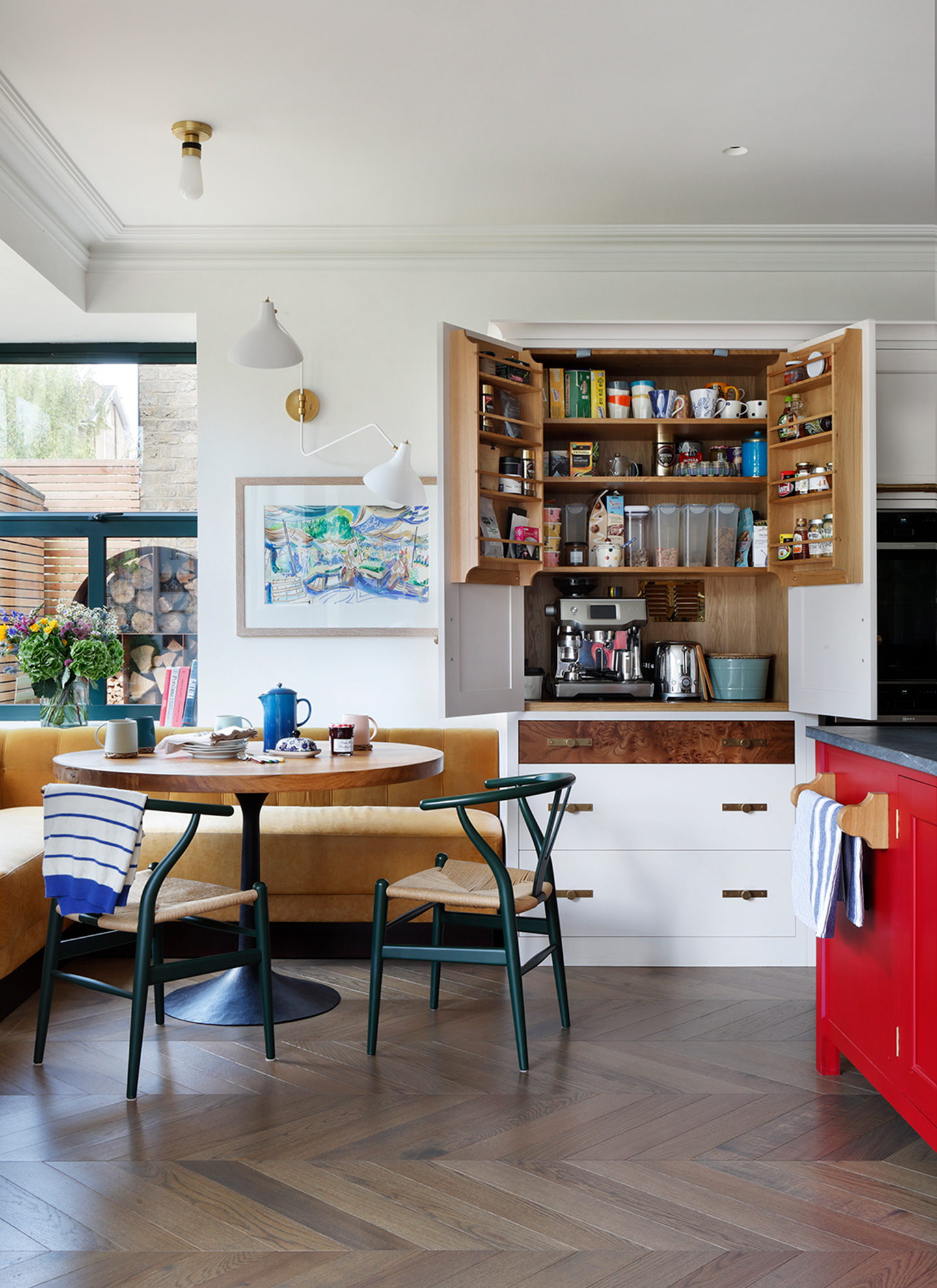 Open kitchen breakfast larder unit with burr wood drawer accents beside a round dining table with mustard banquette seating, showing an espresso machine and organized shelves