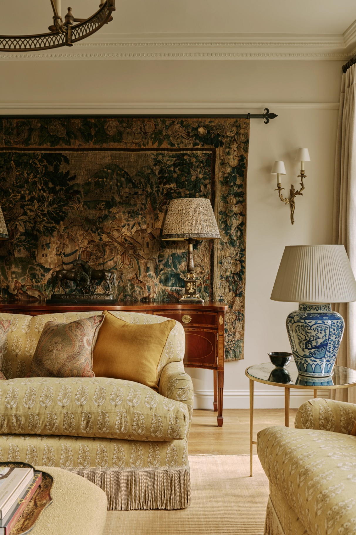 A sitting room with a pair of fringed upholstered classic sofas in the foreground, with a circular side table topped with a large table lamp. In the background is a wooden sideboard and a large classic wall tapestry hanging