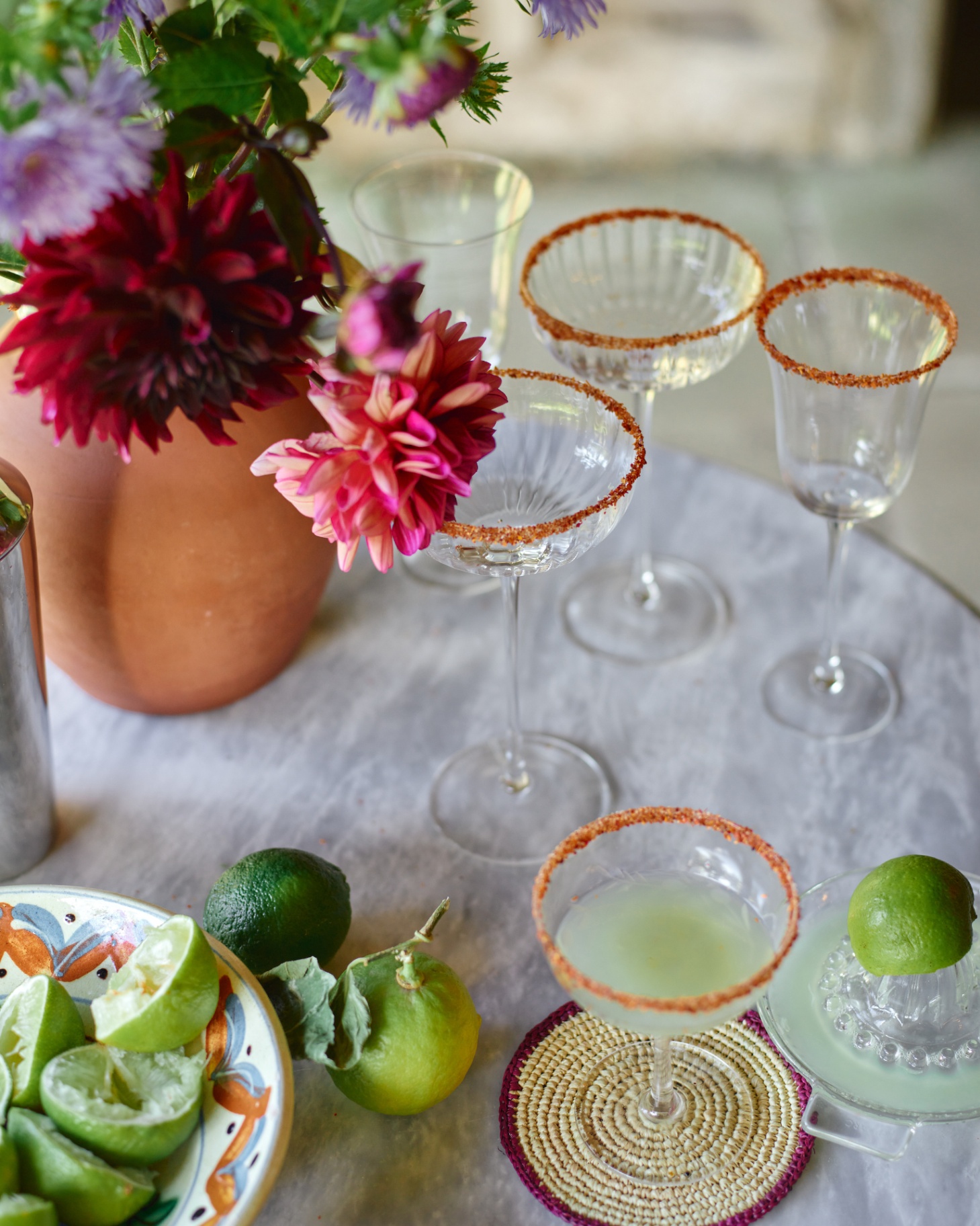 a drink table with 5 glasses with orange salted rims, a bowl of squeezed lemons and a vase of flowers