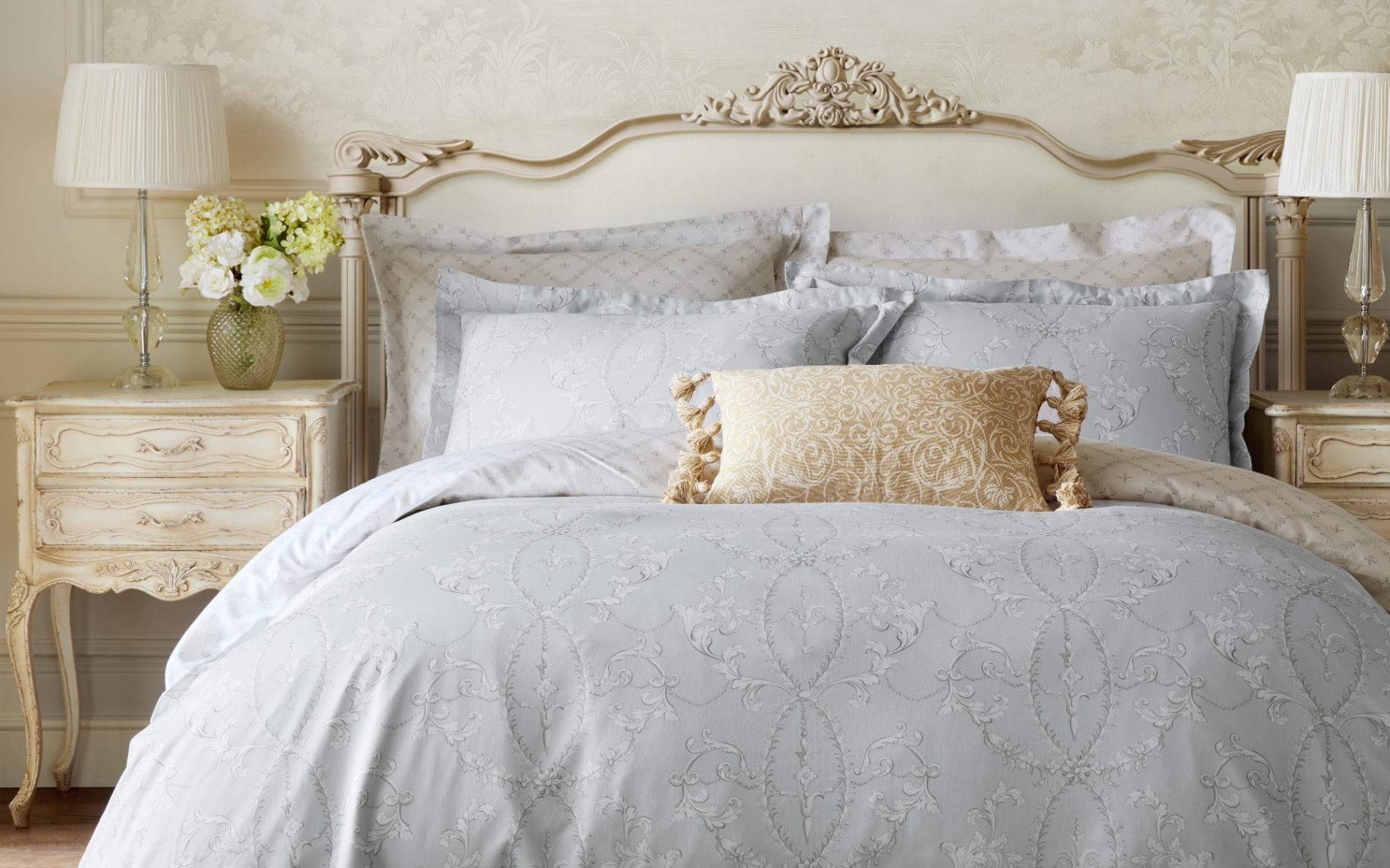 a bedroom with a bed with a cream ornate headboard, grey floral bedding, a yellow patterned cushion and two regency bedside tables with pleated lamps