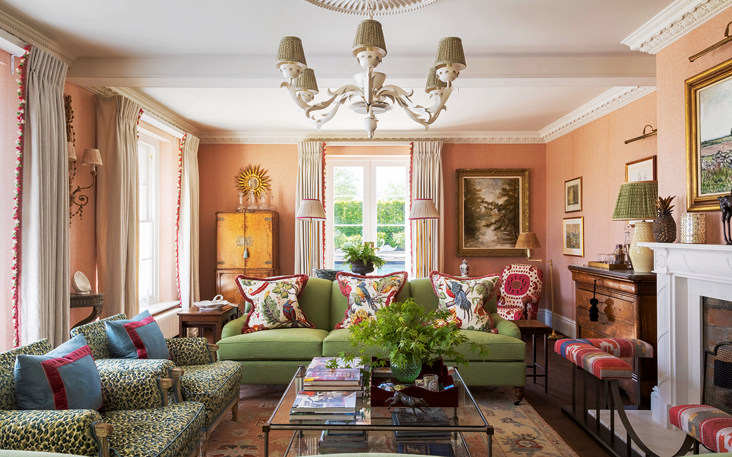 A formal living room with coral walls. Two green-blue leopard armchairs and two green sofas all dressed with decorative cushions surround a glass coffee table topped with a vase of greenery and books. A chandelier hangs above