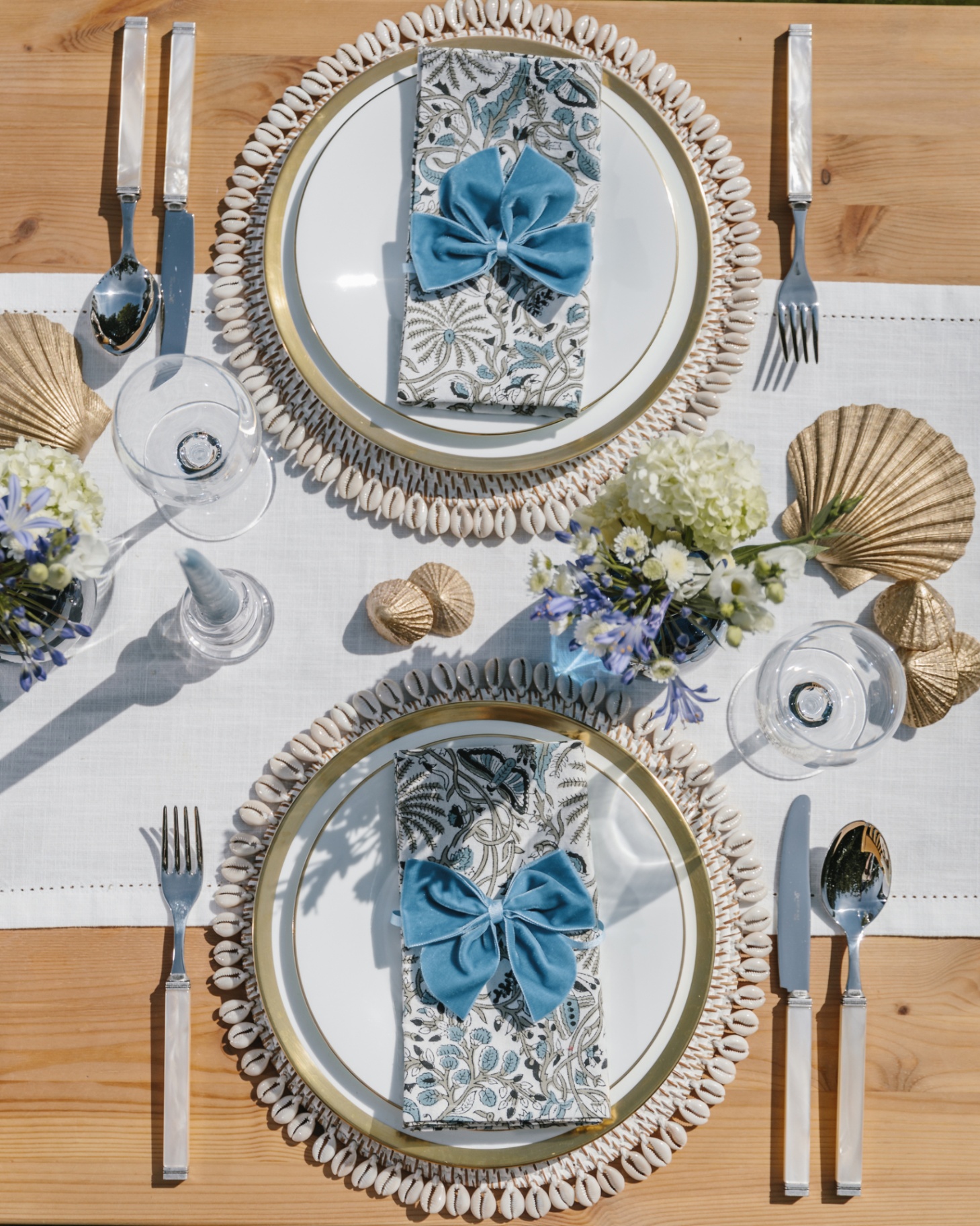 a tablescape with a white runner, circle table mats with a shell beaded trim, napkins on plates with blue velvet ribbon, a small vase of flowers and real seas shells on the table