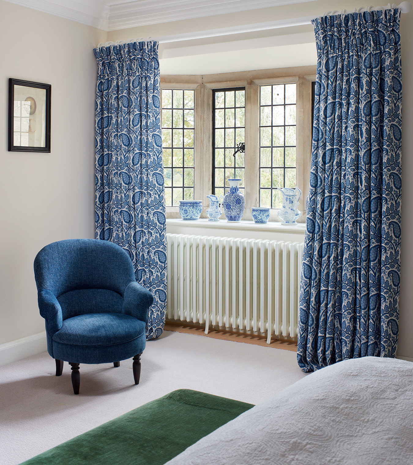 a bedroom corner with a window with blue and white patterned vases on the window sill, blue and white curtains, a blue armchair and a dark green rug