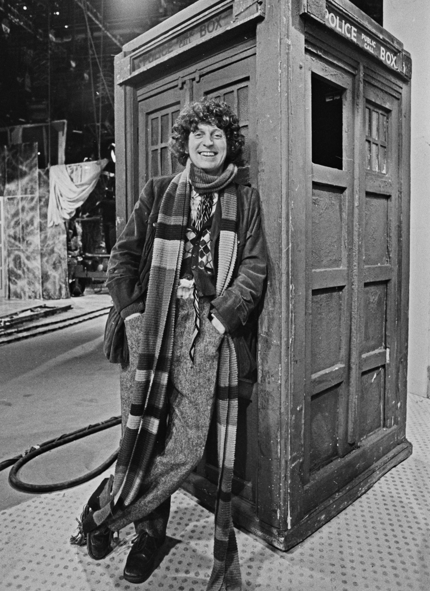 a black and white photo of tom baker standing in front of the tardis in 1974, wearing a long coat and a check scarf