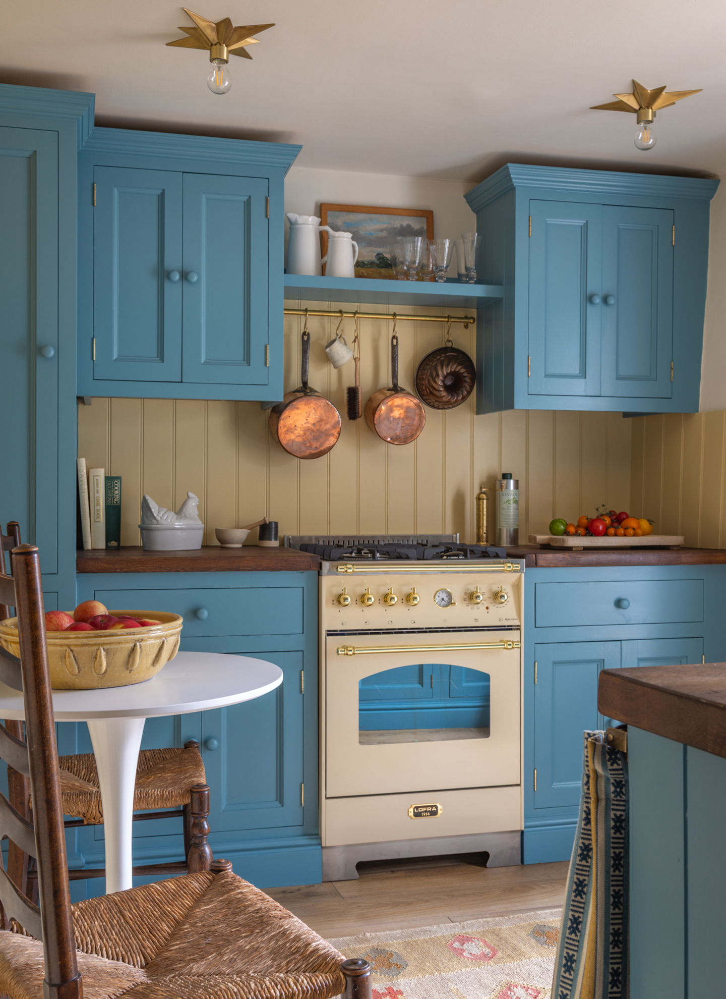 a country kitchen with blue cabinetry, a cream range cooker, two star lights on the ceiling and a shelf with copper pots and pans hanging underneath