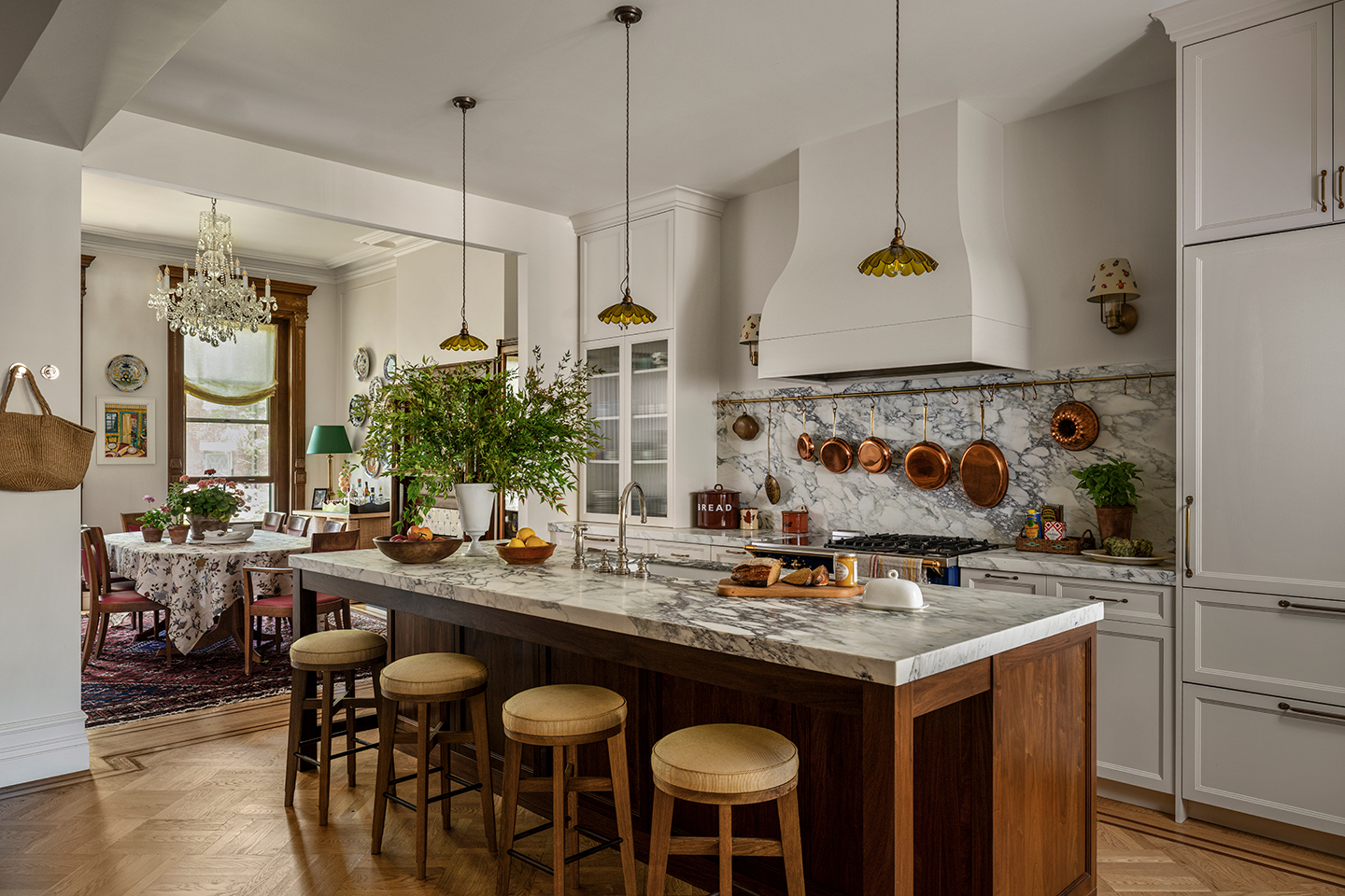 A kitchen with a dark wood and marble topped island in the centre, with three small glass pendants hanging above and bar stools lining the side. White cabinets are paired with marble worktops and blue range cooker. An adjoining dining room is visible 