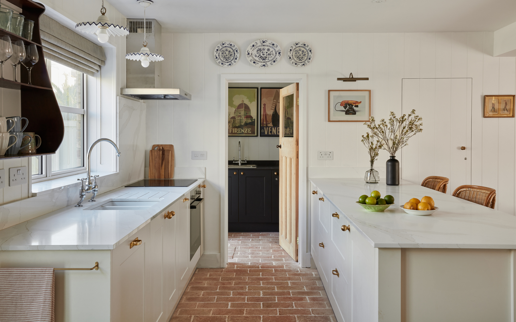 a white kitchen with a brick tile floor, white kitchen cabinets with white marble tops and 3 decorative plates hung above an open door