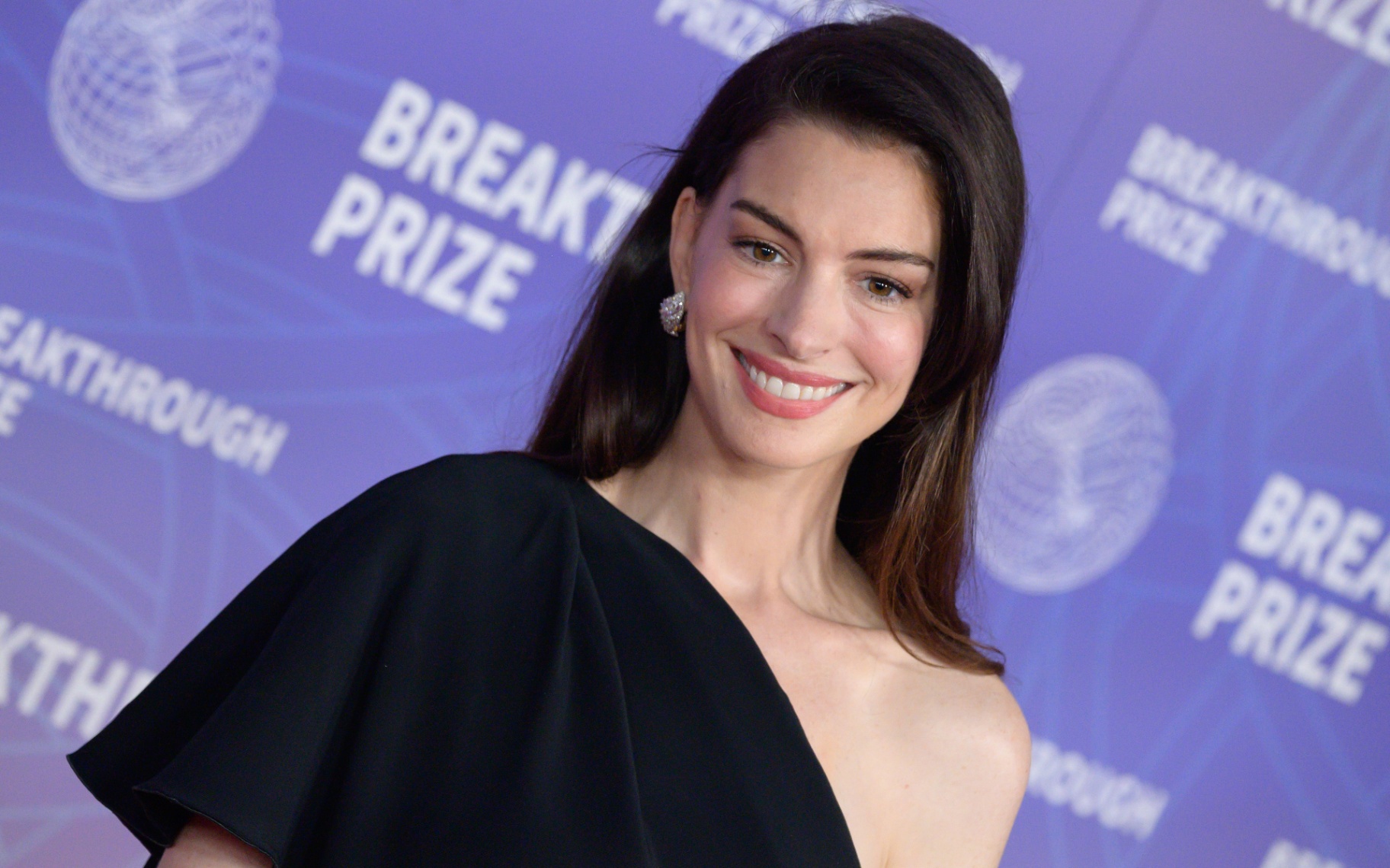 Anne Hathaway wearing a black off the shoulder dress against a bright purple backdrop