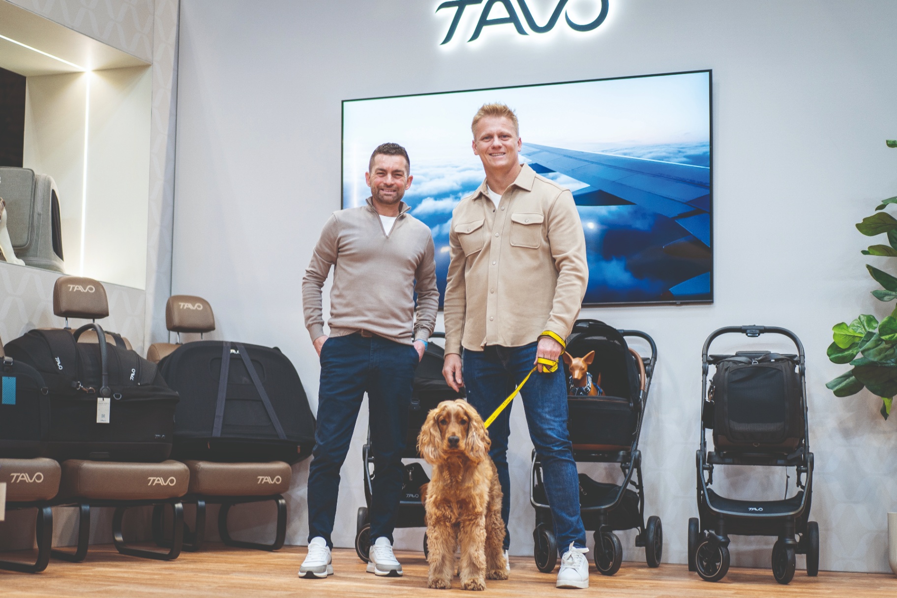 TV vet Dr Scott Miller and dog behaviourist Adem Fehmi standing with a golden cocker spaniel at a Tavo exhibition display showcasing pet car seats and travel carriers