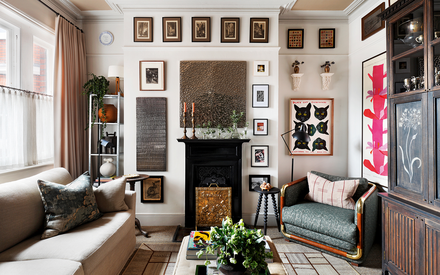 a small living room with a neutral colour scheme. A sofa sits on the left below a window opposite a mid-century armchair and bespoke painted dark wood cabinet. In the centre of the image is a narrow fireplace, with a large collection of artwork above