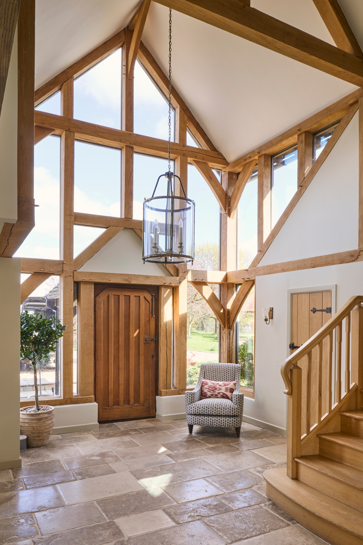 A large entrance hall with a vaulted ceiling made from wood and windows, with limestone flooring and a large lantern pendant hanging in the middle. An armchair sits in the corner and in the foreground are some wooden stairs 