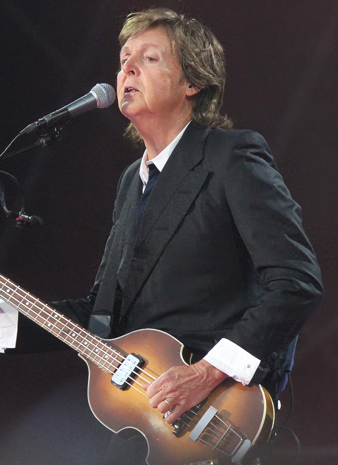 paul mcCartney wearing a black blazer singing into a microphone and playing the guitar on stage