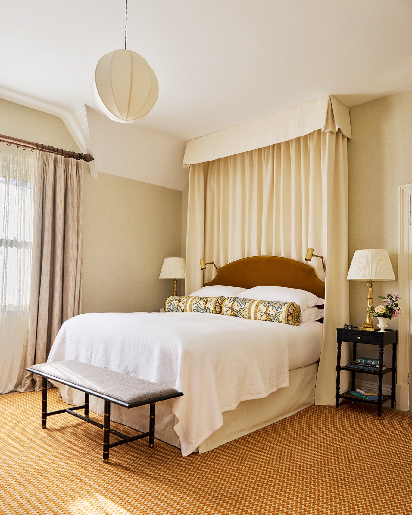 A neutral bedroom with a double bed with a dark yellow velvet headboard, a cream fabric canopy hanging above, and dressed with white bed linen and patterned bolster cushions. In the foreground at the end of the bed is a bench with an upholstered seat. 