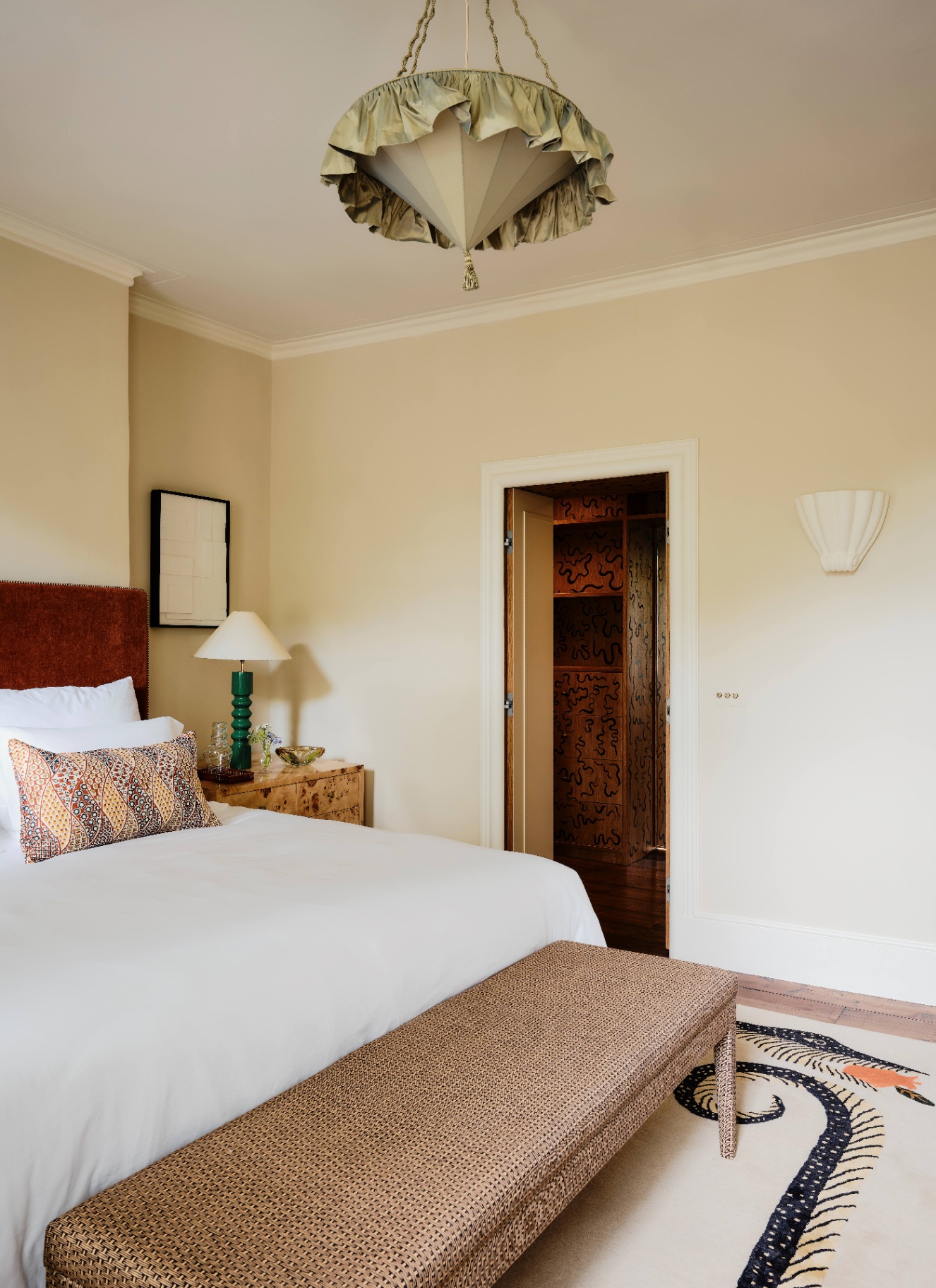 A neutral master bedroom with a green stacked-base bedside lamp featuring a white conical lampshade beside a brown velvet headboard