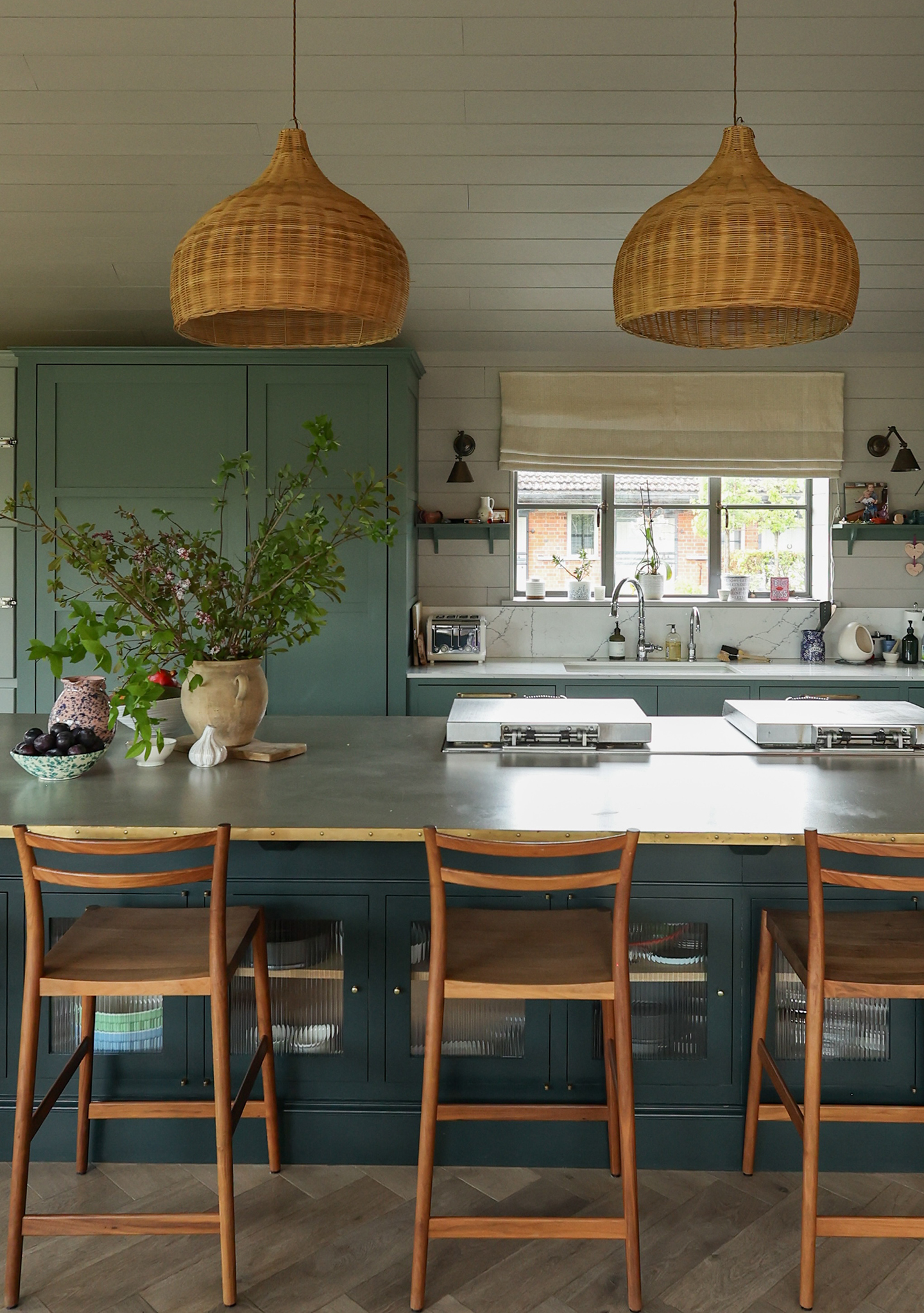 A large kitchen island and wooden bar stools sit in the foreground, with large rattan pendants hanging overhead. In the background are more cabinets, painted blue, and a sink area with a window dressed with a blind above