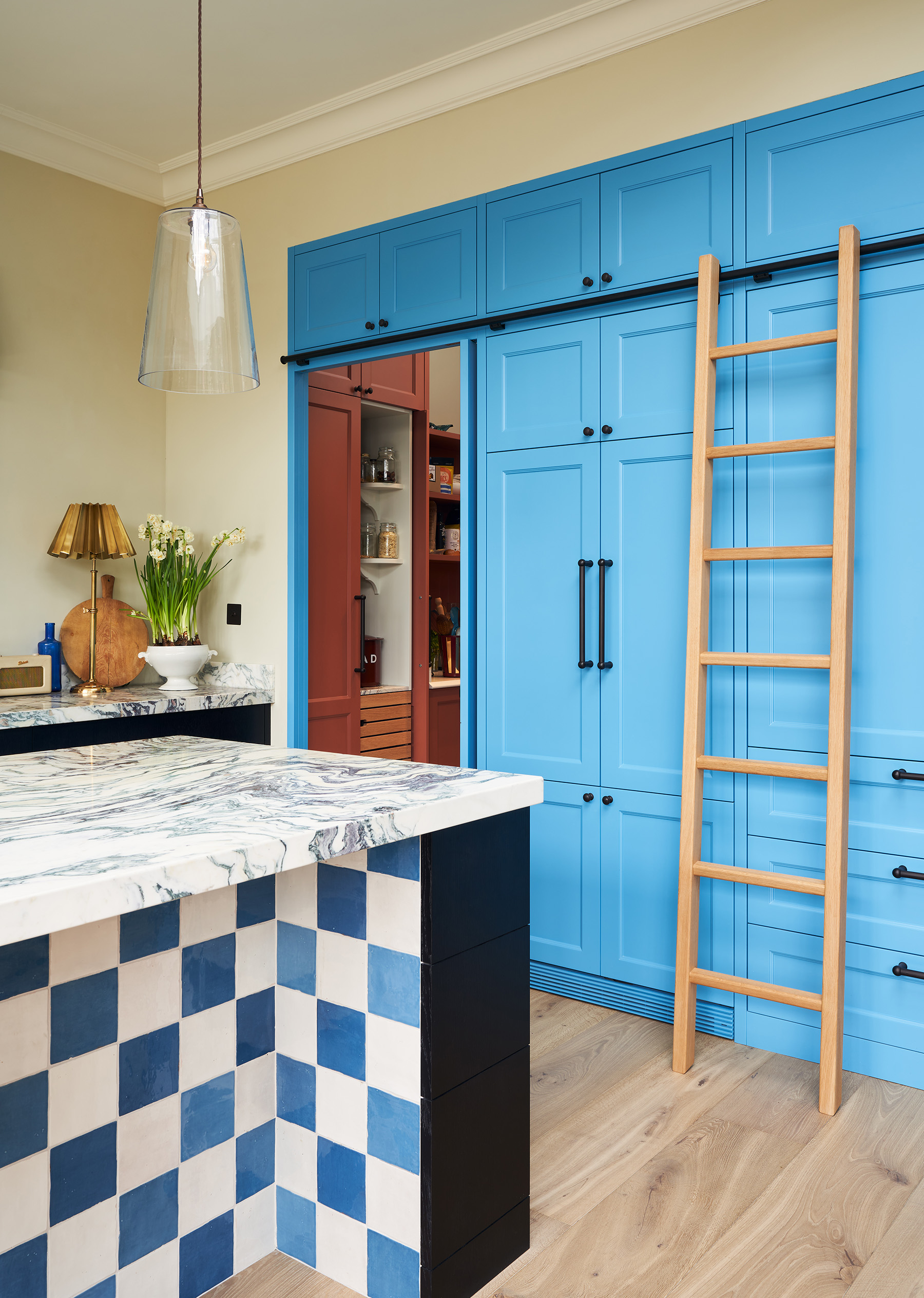 a pantry door set in a row of tall, built-in kitchen cabinets finished in a vibrant shade of blue and with an upper rail which has a ladder to run along it and in the foreground is a tiled kitchen island in white and blue and black with marble top