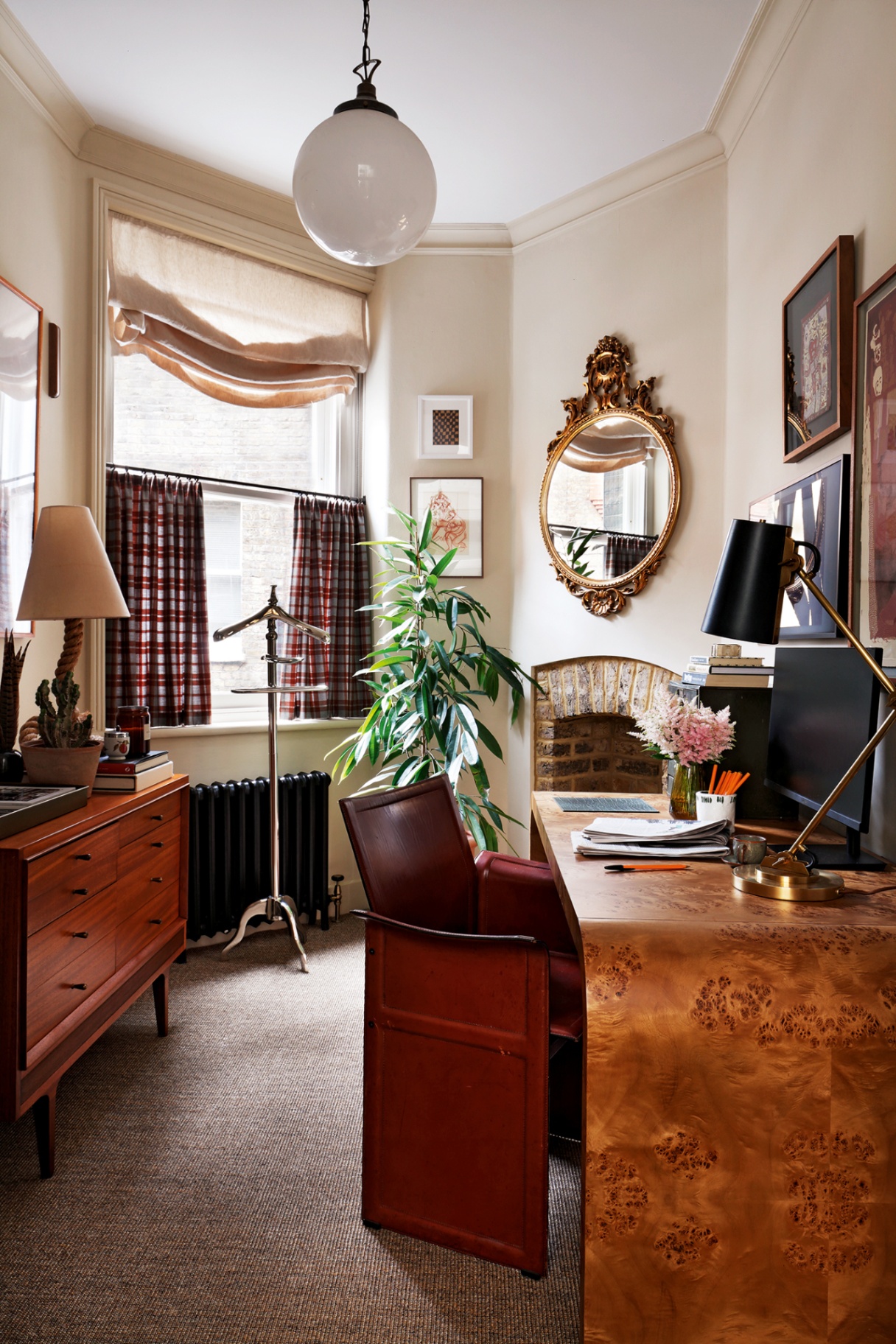 A home office with a burl wood desk in the foreground, with a red leather desk chair and topped with stationery, computer and table lamp. A globe pendant hangs above while a wooden sideboard lines the left wall. An antique mirror and art hangs on the walls