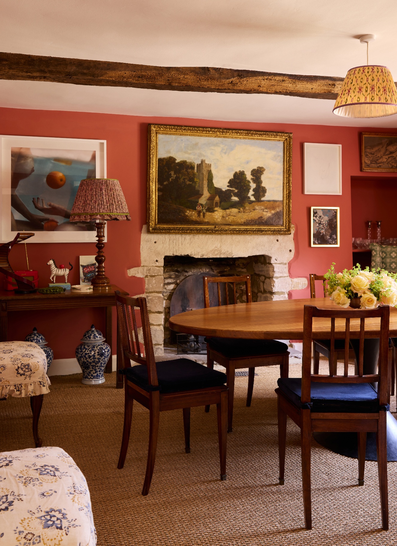 a dining room with coral orange walls, wooden beams on the ceiling, a large stone fireplace with a large lanscape painting above it, a long wooden table with dark wooden dining chairs and a jute carpet