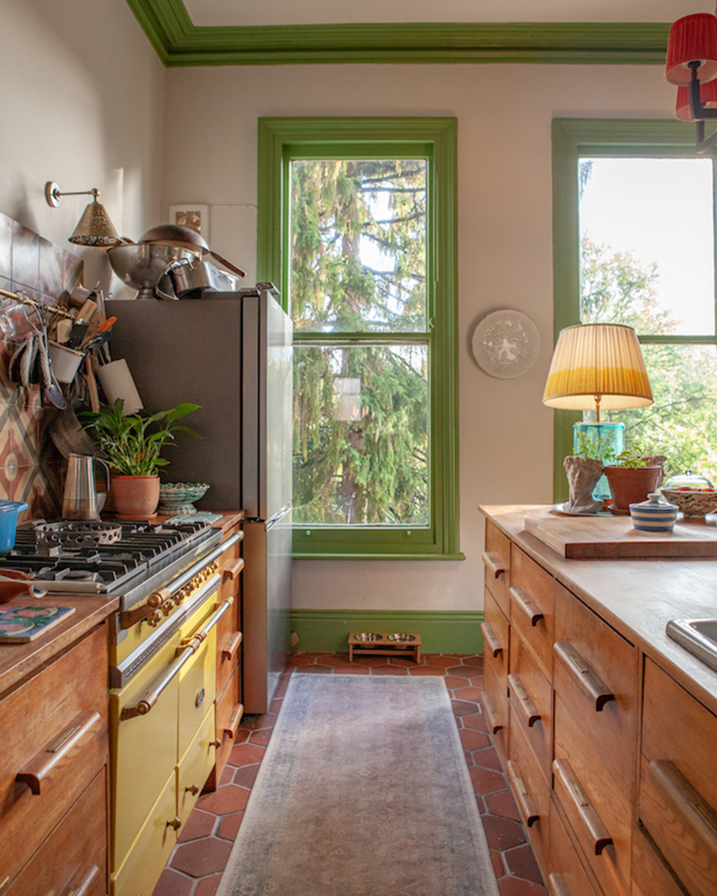 a country kitchen with brick flooring, green painted joinery, a yellow range cooker and a messy countertop, and a table lamp on the opposite counter 