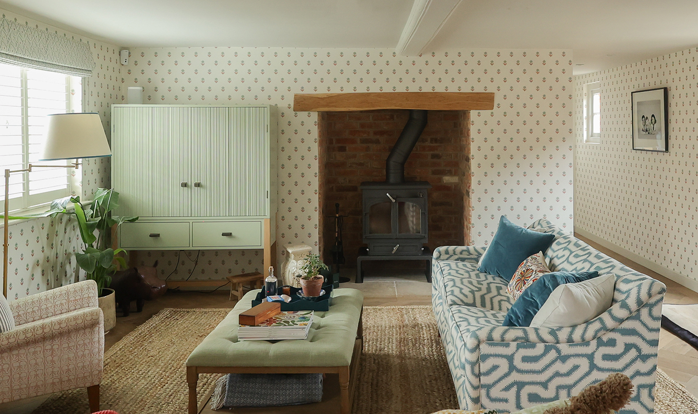 A snug lined with a dainty wallpaper. On the back wall is a woodburner in the fireplace and a green ribbed cabinet. In the foreground, a patterned sofa and armchair sit either side of an upholstered coffee table, all arranged on a jute rug