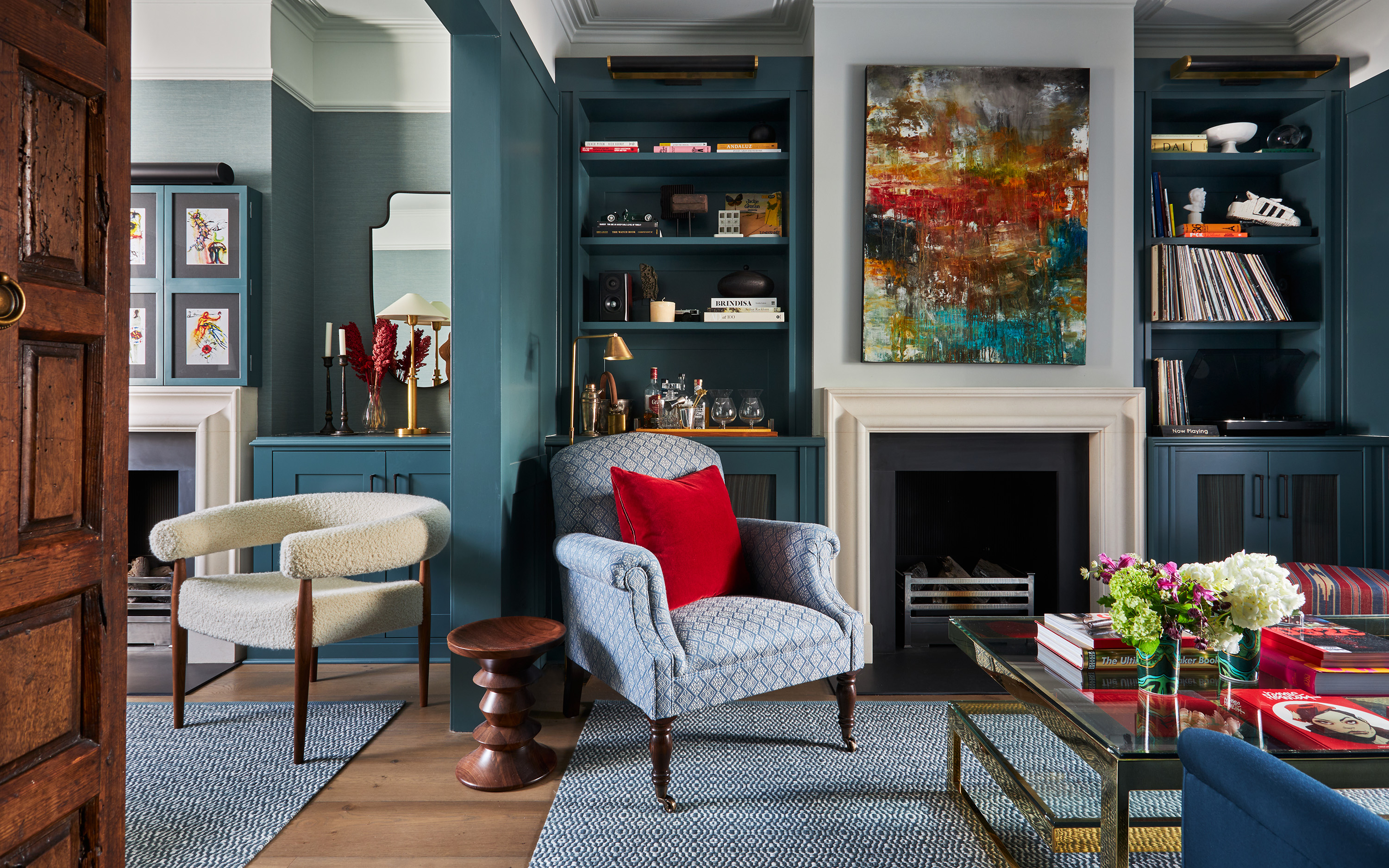a living room with dark navy walls with built in bookcases, a alrge abstract art canvas above the fireplace and a blue and white patterned armchair with a red cushion