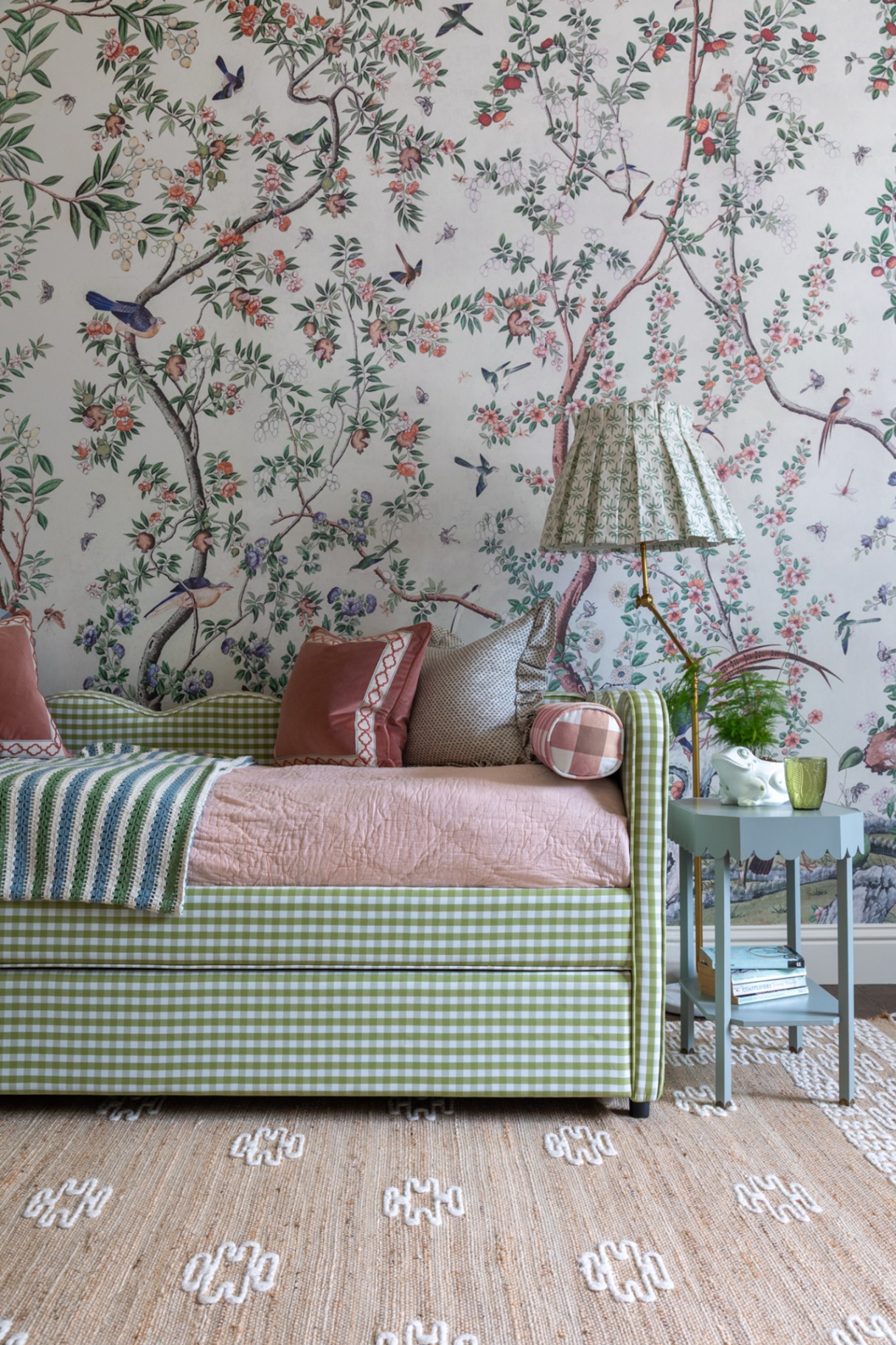 A green-and-white gingham daybed styled with pink and patterned cushions set against botanical floral wallpaper. Beside it sits a blue accent table with a brass floor lamp and a natural woven area rug