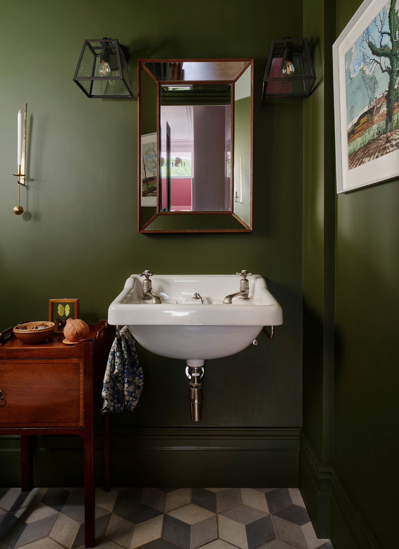 Olive green bathroom featuring a wall-mounted white ceramic basin with exposed chrome plumbing and a copper-framed mirror overhead. Black lantern sconces flank the mirror against painted green walls, set above geometric floor tiles and a wooden table