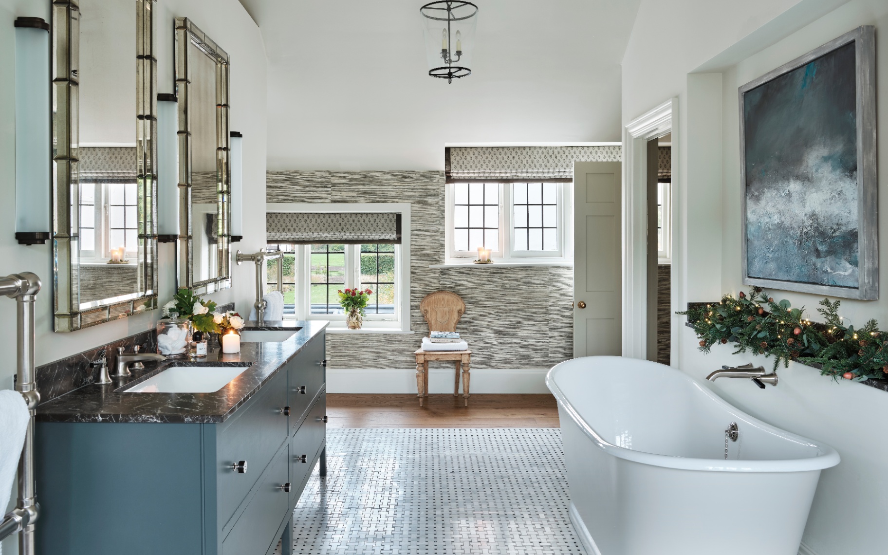 An elegant bathroom featuring a double vanity with a dark countertop on the left and a freestanding white bathtub on the right. A festive Christmas garland with fairy lights is draped on a shelf above the tub. Large windows and a tiled floor.