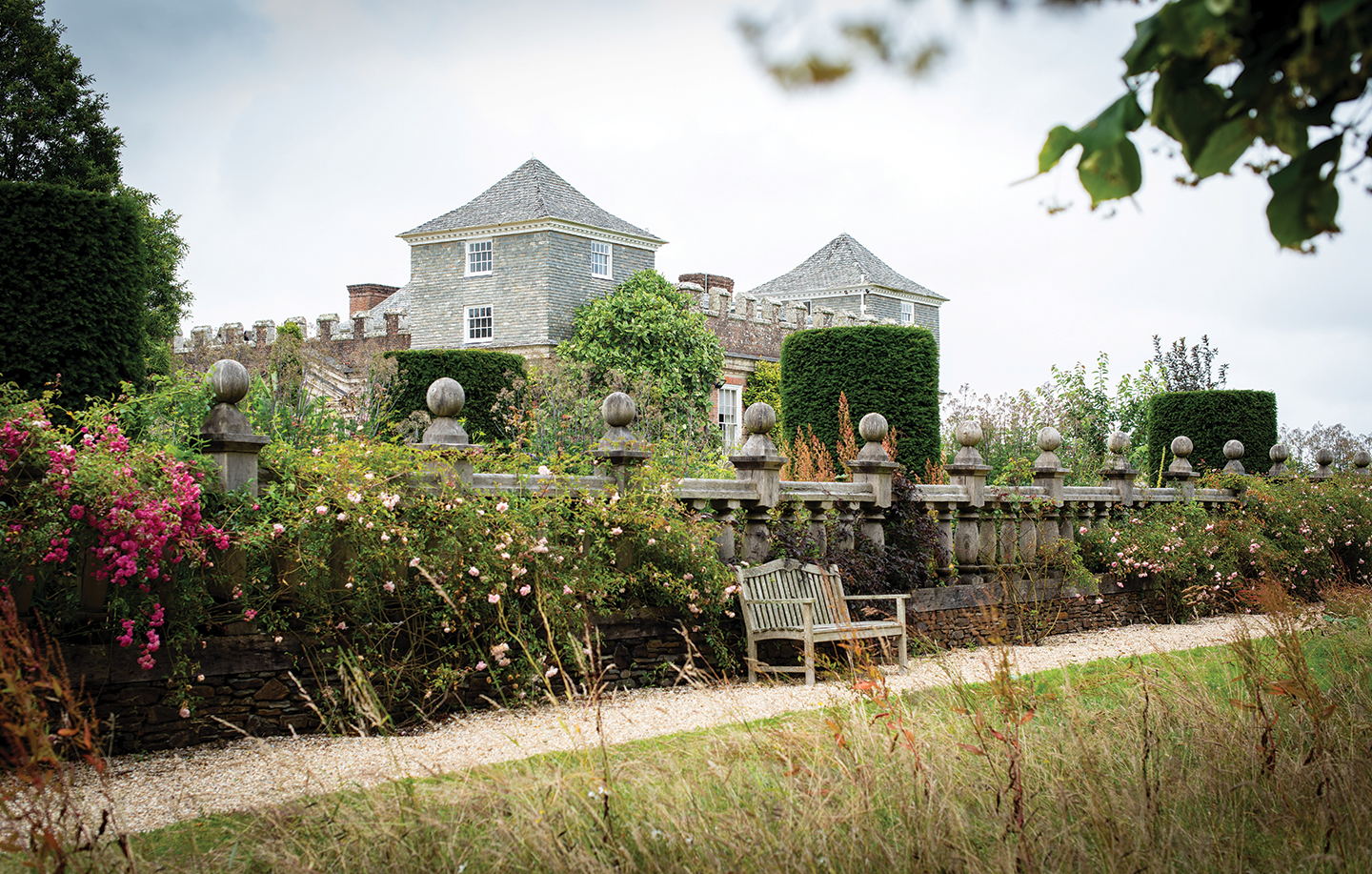 an exterior shot of a 17th century manor house with battlemented towers partly obscured by a stone wall with flowers and a bench in front.