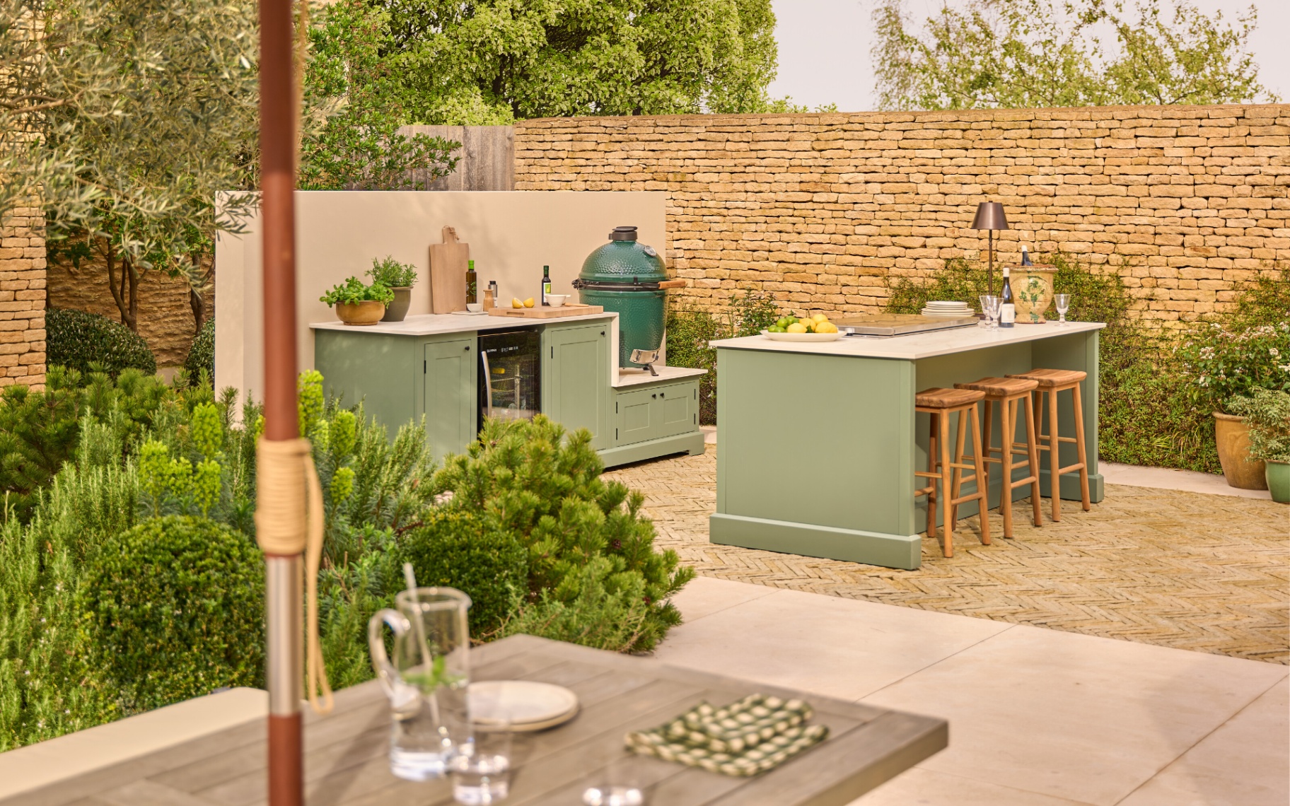 a sage green outdoor kitchen in front of a cotswold stone brick wall with trees behind it and a wooden outdoor dining table in the foreground with a glass jug on top