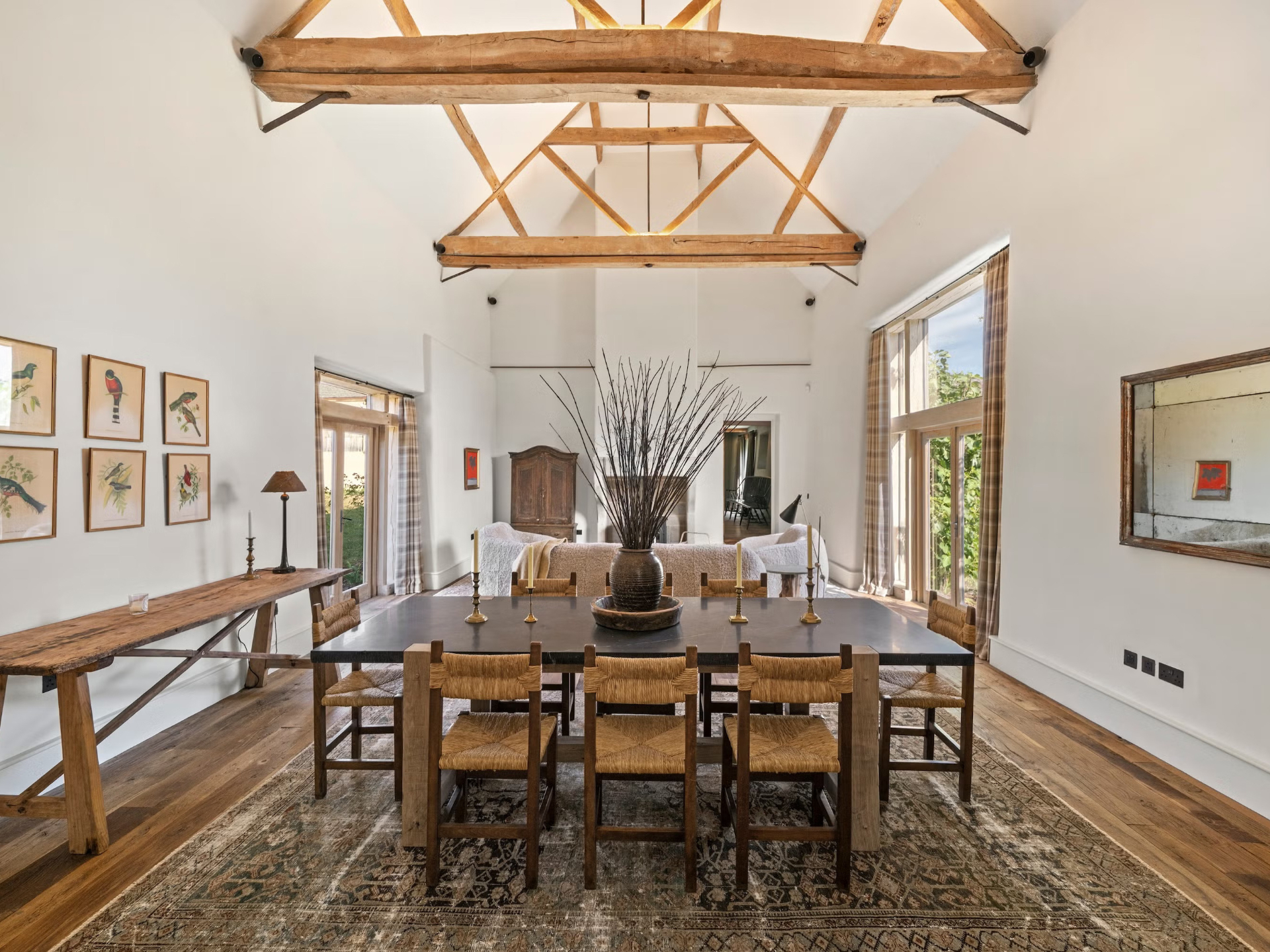 a dining room with tall ceiling and wooden beams, a large dining table with wicker seats and a wall covered in prints of birds