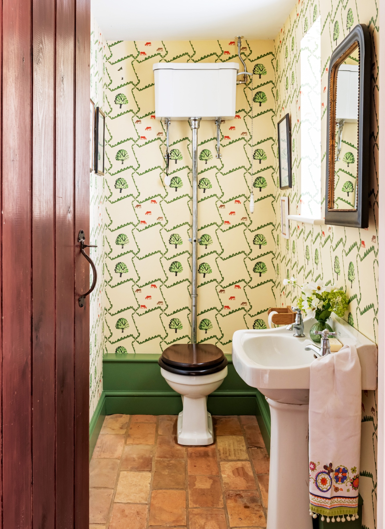 a downstairs toilet with a cream and green botanical print wallpaper, a high level cistern, a white sink with a white patterned towel on it and a black arched mirror above