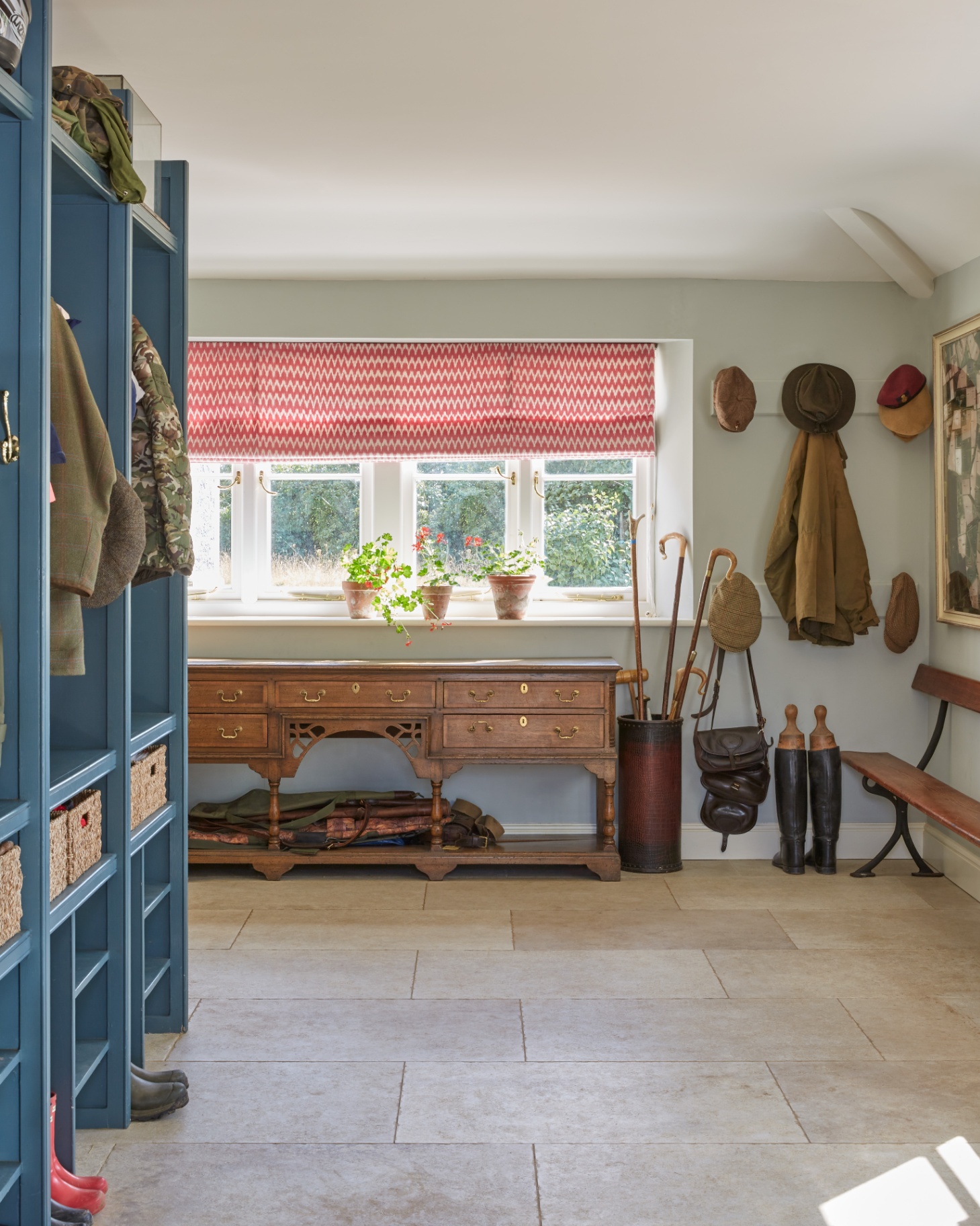 a boot room with pale stone floors, a red and white patterned blind on a window, a blue painted shelving unit to the left and boots, coats and hats hung on the back wall